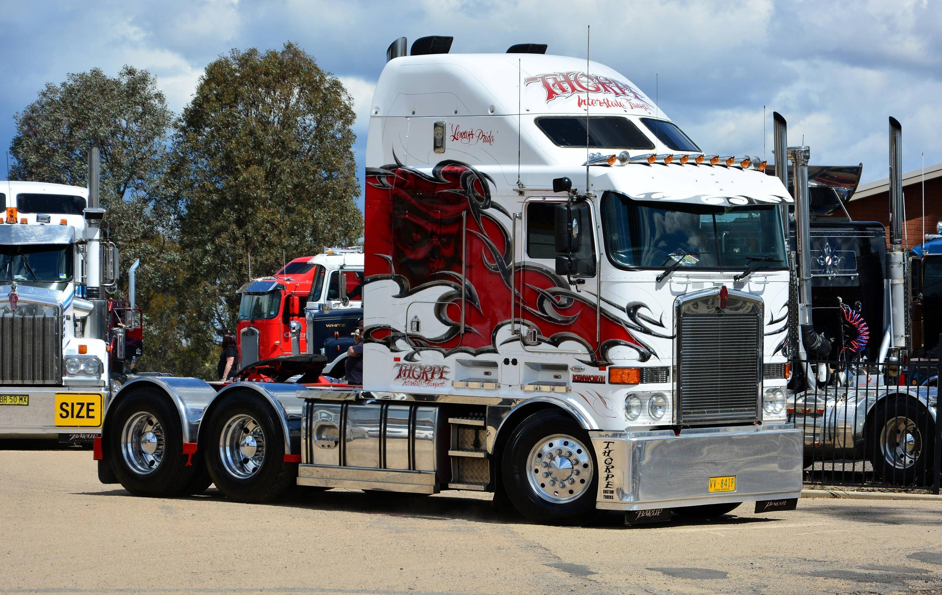 Kenworth客製化紅色骷髏貼紙 壁紙 — 一輛帶有紅色火焰貼花和鍍鉻裝飾的白色半掛卡車，停放在陽光明媚的停車場，周圍有其他卡車和樹木。