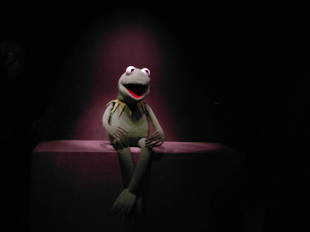 Kermit 舞台聚光燈 壁紙 — 一盞聚光燈照亮了坐在黑暗劇院基座上的青蛙柯密特，他睜大的眼睛和張開的嘴巴表達了驚訝或喜悅。