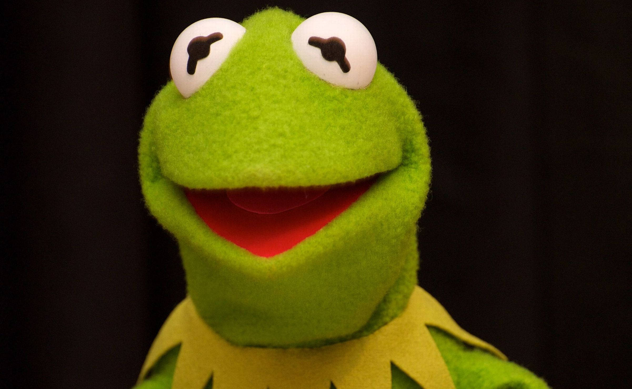 Kermit 黑底圖像 壁紙 — 青蛙柯密特布偶的特寫，帶有亮綠色氈質感和寬闊歡樂的笑容，背景為深色。