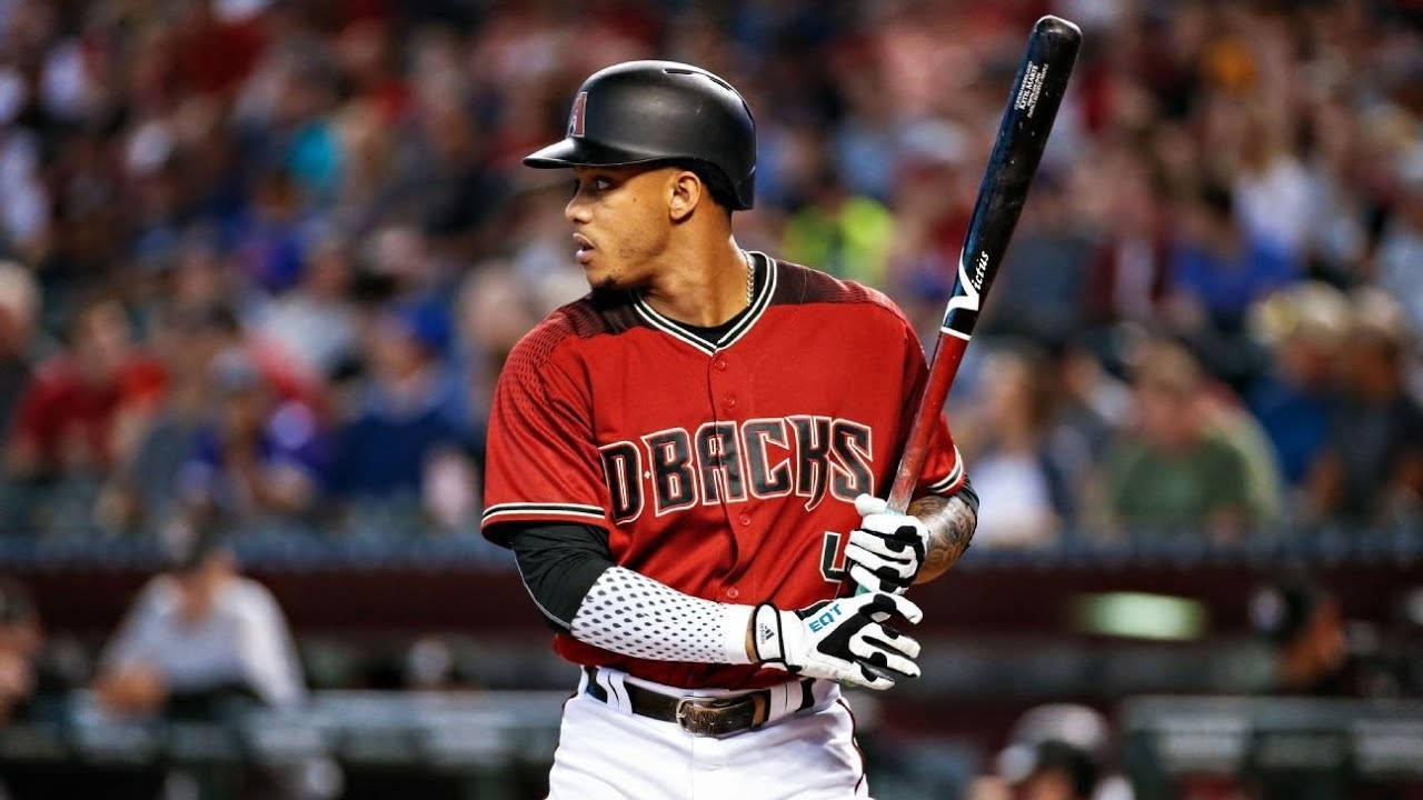 Ketel Marte 在場上專注比賽 壁紙 — 一名專注的棒球運動員身穿紅色DBacks制服，在比賽中緊握球棒，身後是模糊的體育場人群。