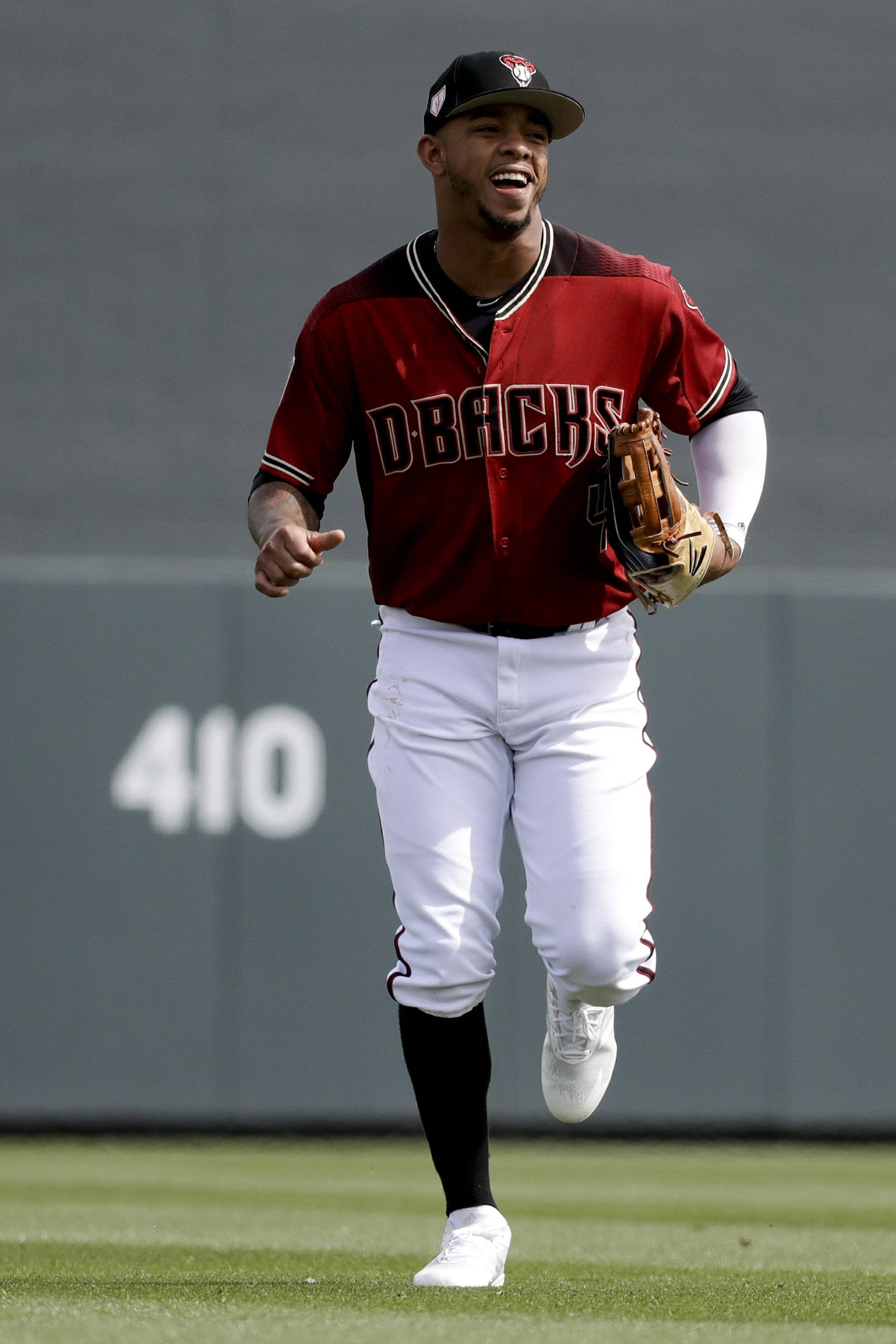Ketel Marte 跑步 壁紙 — 一位微笑的棒球運動員身穿紅色Dbacks球衣，戴著手套在綠色場地上快樂奔跑。