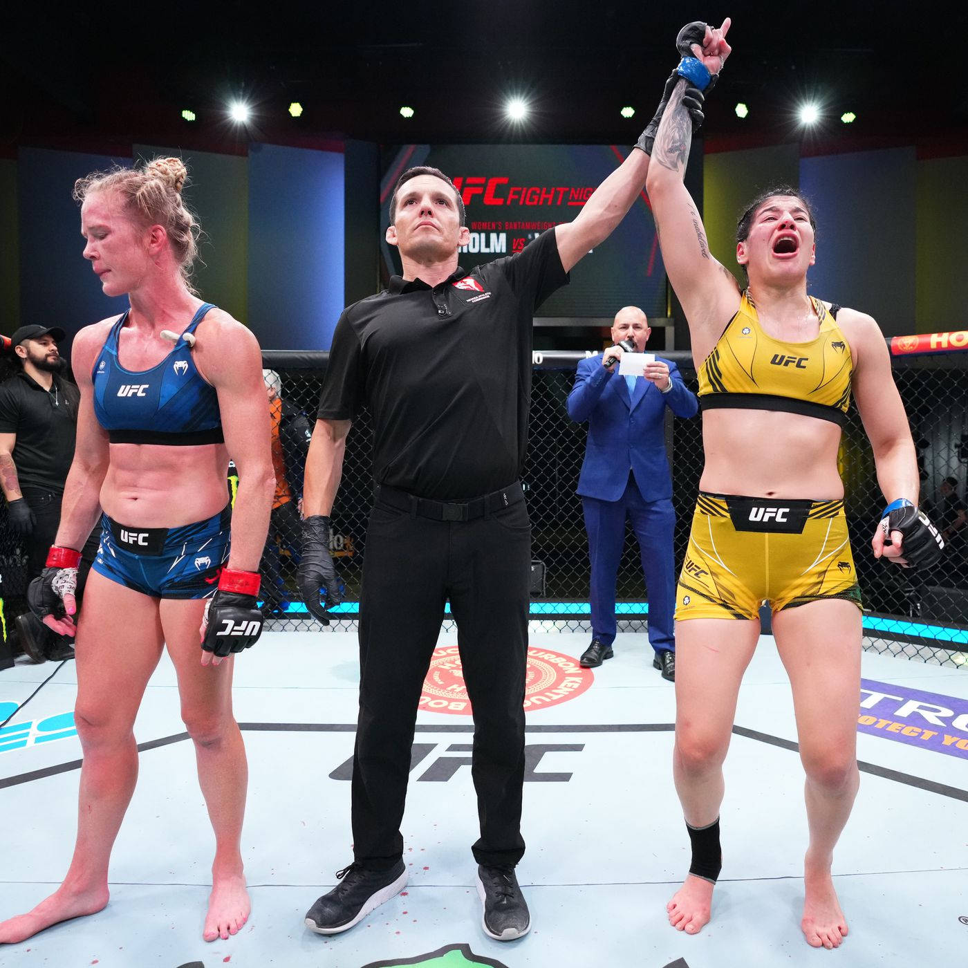 Ketlen Vieira 擊敗 Holly Holm 壁紙 — UFC裁判舉起身穿黃色裝備的勝利拳手，而身穿藍色裝備的對手在八角籠中觀望。