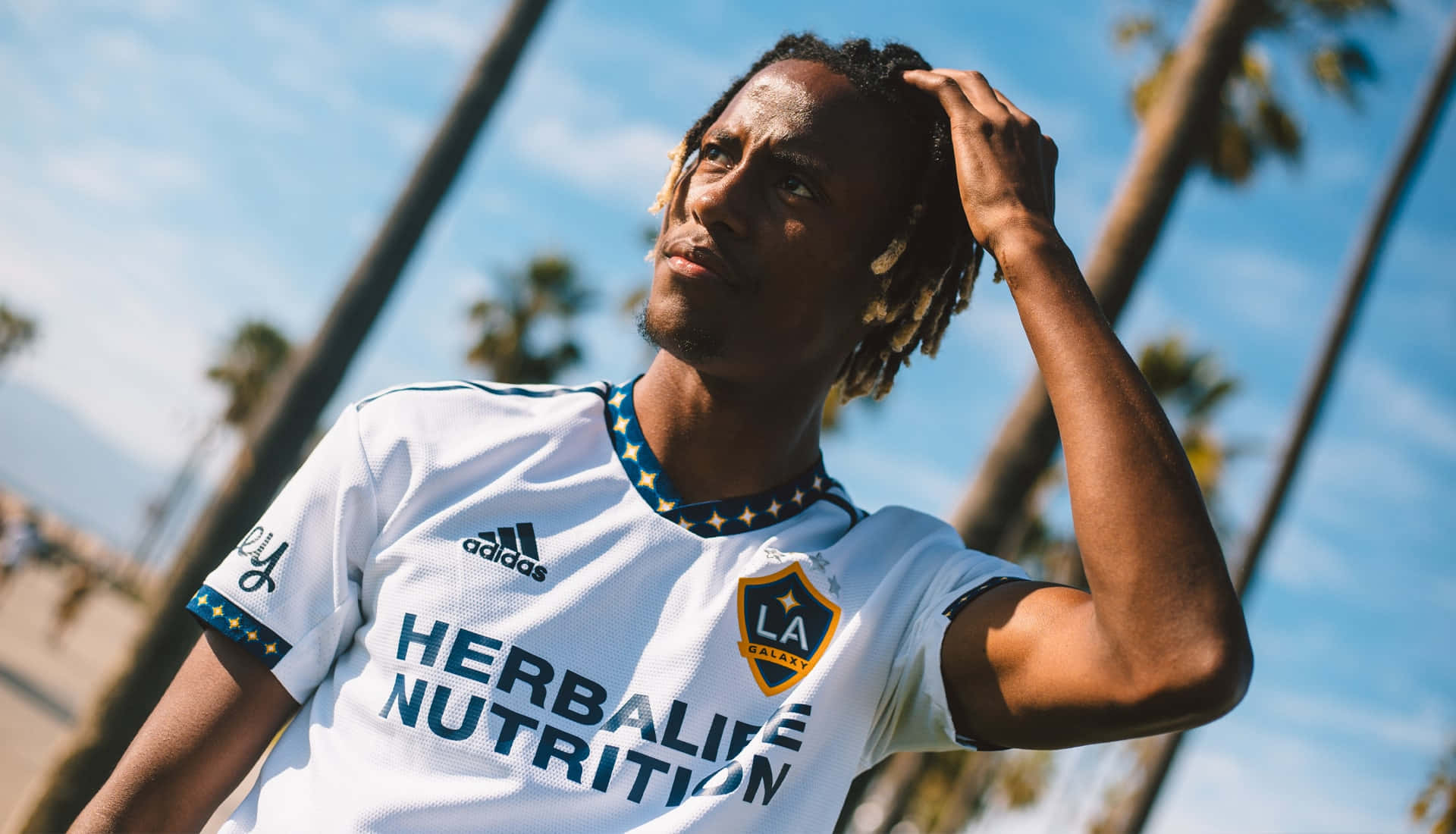 Kevin Cabral 穿 LA Galaxy 2022 Adidas 主場球衣 壁紙 — 一名身穿白色洛杉磯銀河球衣的足球運動員，在藍天白雲和棕櫚樹背景下自信地站立。
