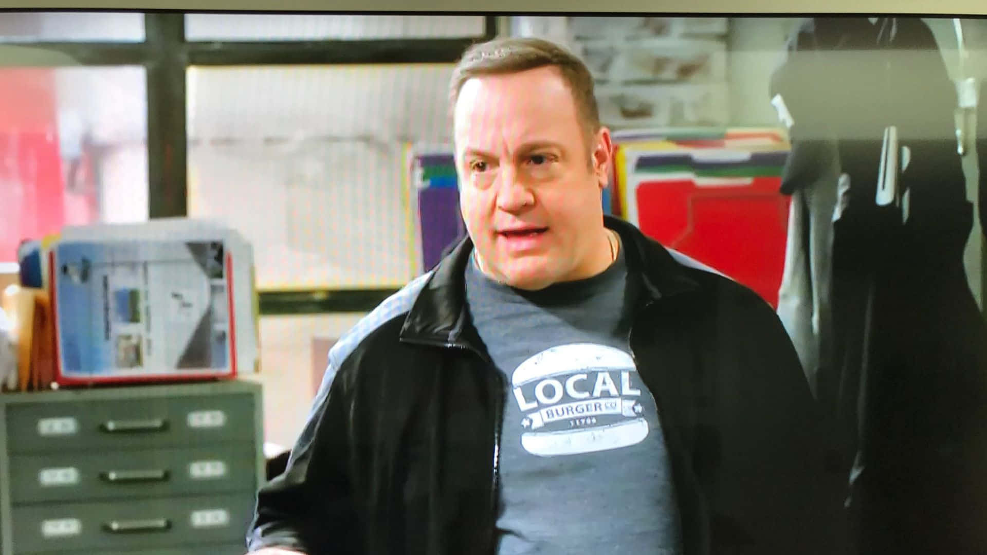 Kevin James 休閒造型 壁紙 — 一名身穿黑色外套和灰色「Local Burger Co.」T恤的男子站在雜亂的辦公室裡，身後有文件櫃和彩色文件夾。