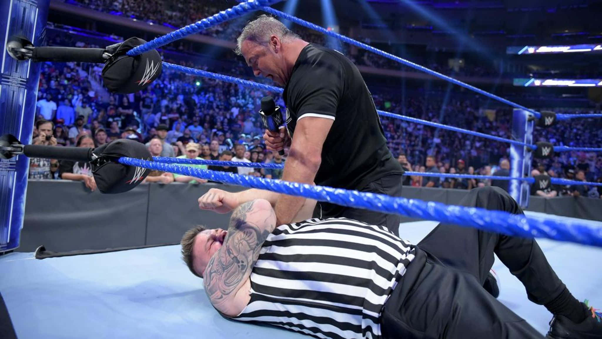 Kevin Owens 對決 Shane McMahon WWE Smackdown 壁紙 — 一名身穿黑色襯衫的摔角手攻擊一名身穿條紋襯衫、躺在藍色摔角墊上的裁判，場地燈光明亮。