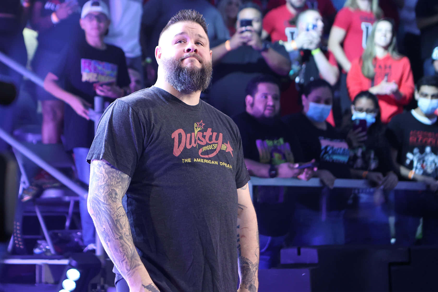 Kevin Owens 穿著 Dusty Rhodes 紀念商品 壁紙 — 一位留鬍子、有紋身的男人自信地站在燈光昏暗的競技場中，粉絲們在看台上觀看。