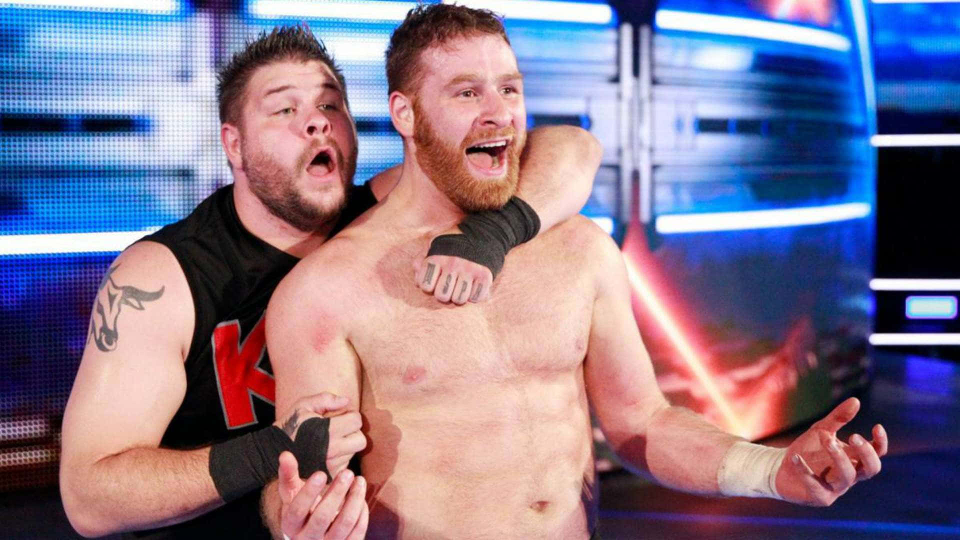 Kevin Owens 與 Sami Zayn WWE 壁紙 — 兩名肌肉發達的摔角手在燈光璀璨的擂台上戲劇性地擁抱，藍色和紅色的燈光交織。
