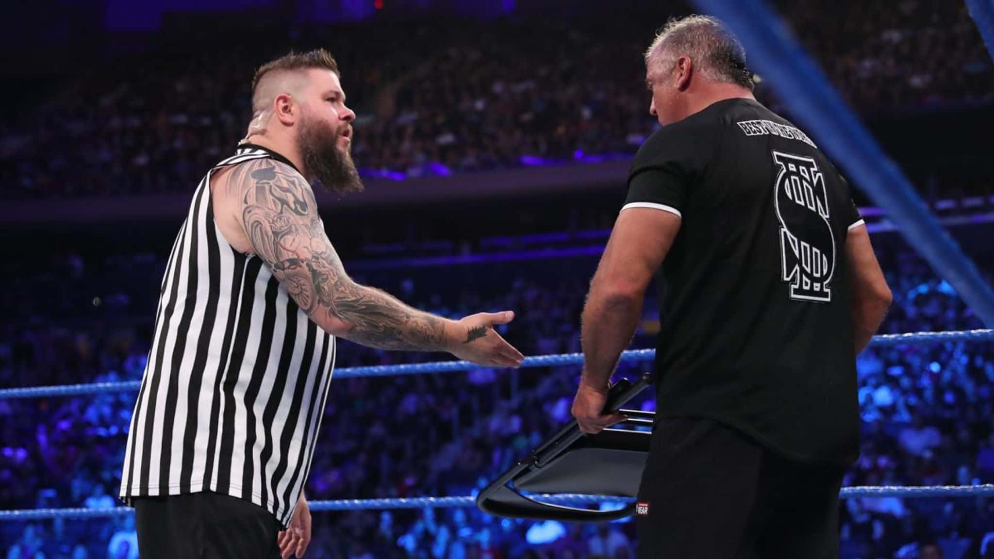 Kevin Owens 與 Shane McMahon WWE 夏季大賽對話 壁紙 — 一位有紋身的裁判穿著條紋衫，在燈光明亮的摔角場內與一名手持椅子的摔角手對峙。