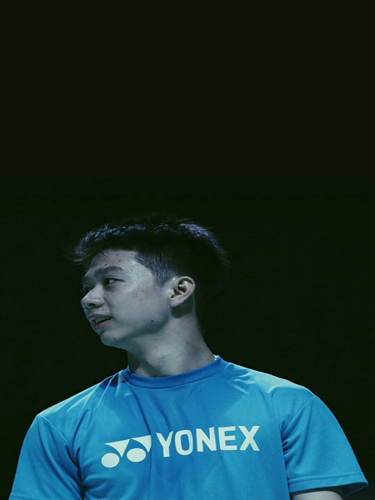 Kevin Sanjaya 搭配Yonex球衣 壁紙 — 一位身穿藍色Yonex球衣的年輕羽毛球運動員在戲劇性燈光下，對著黑暗背景專注地凝視。