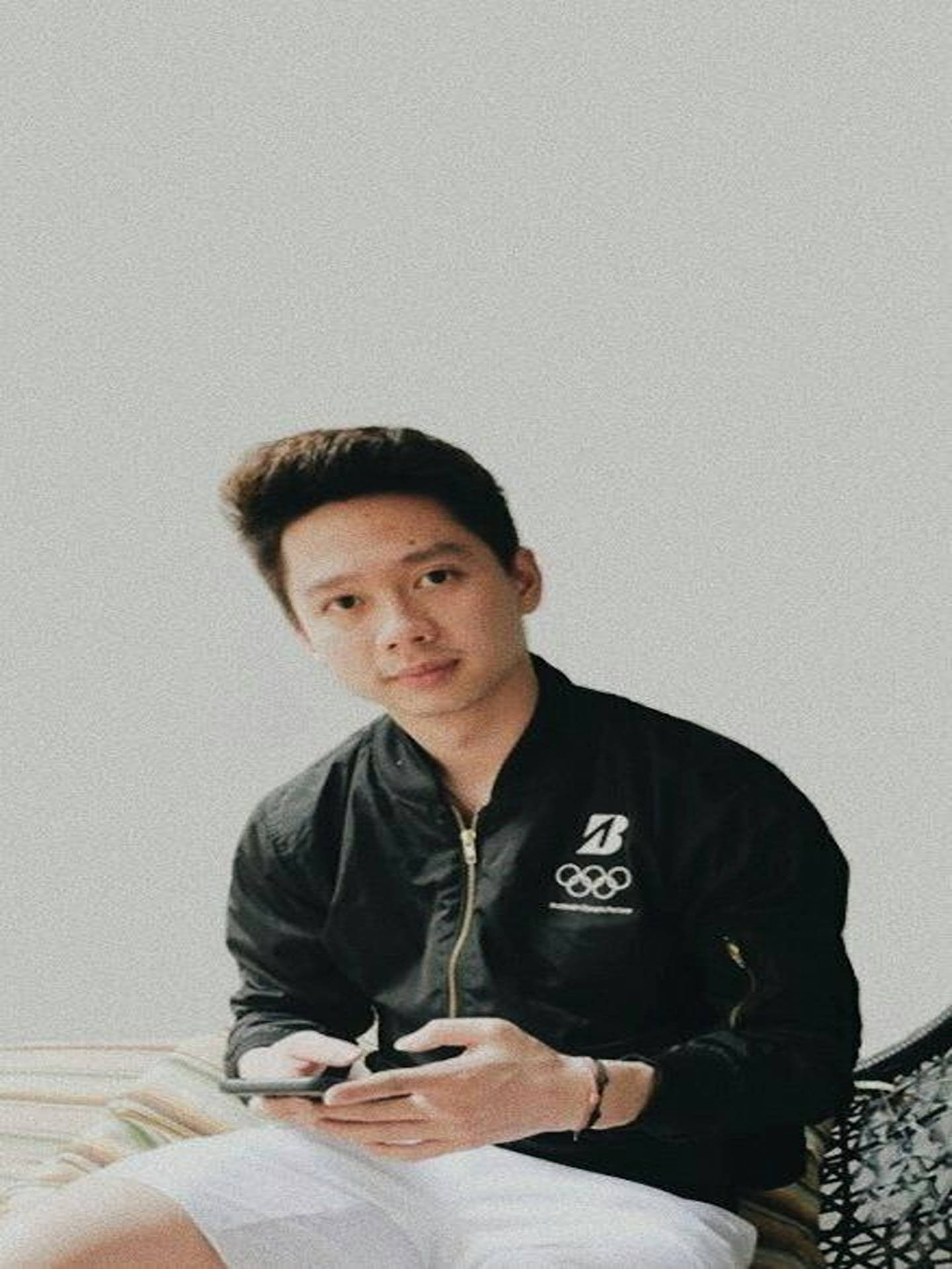 Kevin Sanjaya 玩手機 壁紙 — 一名身穿黑色奧運外套的年輕男子平靜地坐著，手持電話，背景為純灰色。