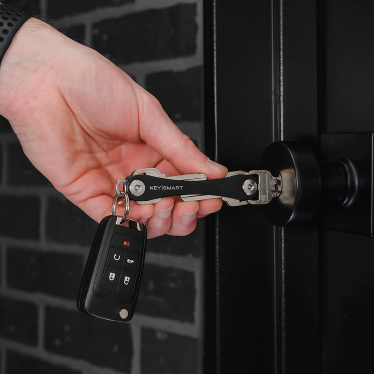 手拿著車匙和KEYSMART整理器，黑色門上，磚牆背景。