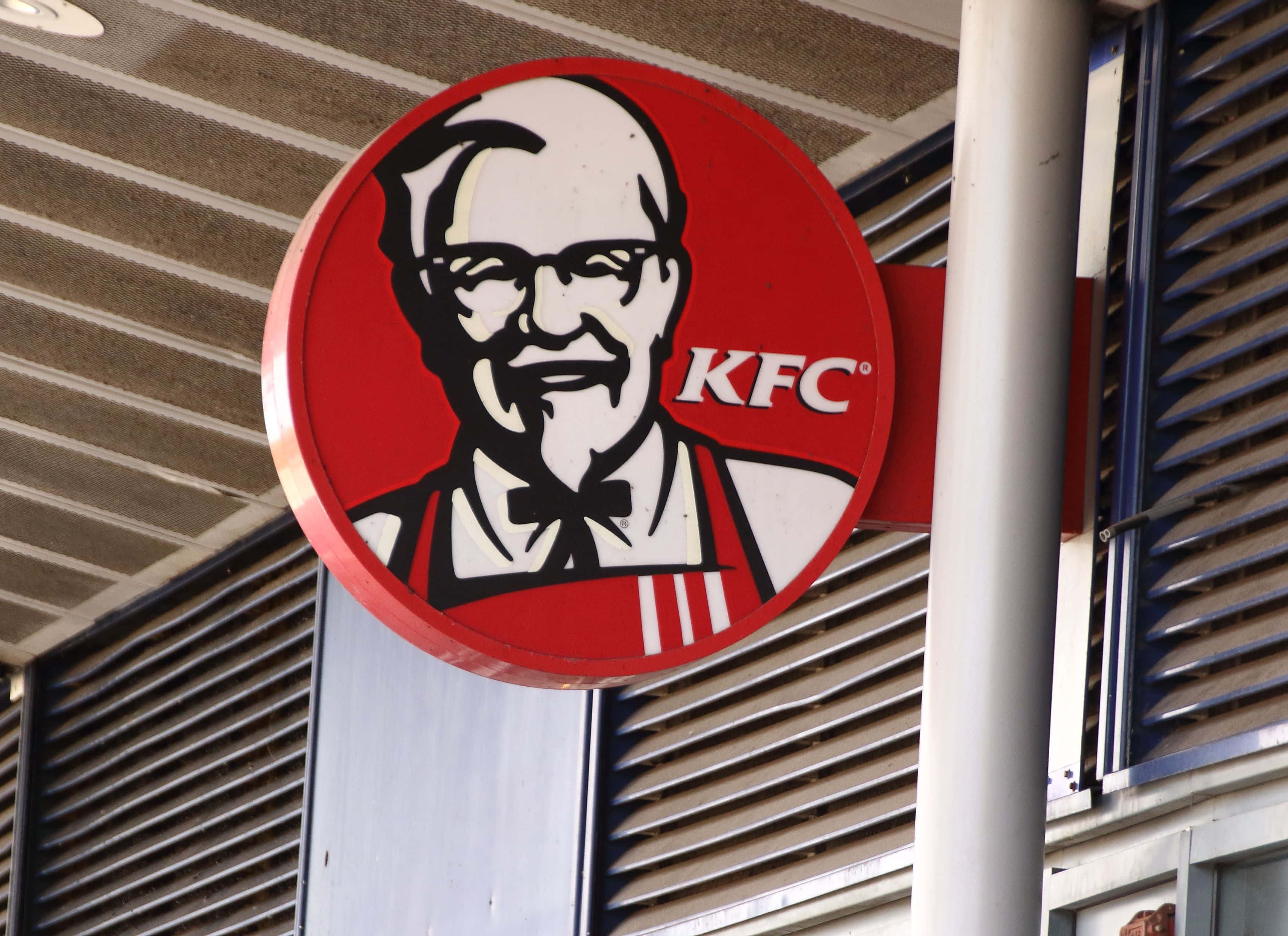 KFC 用心製作 100% 真雞料理 壁紙 — 經典紅色圓形KFC招牌特寫，上面有山德士上校的笑臉，背景是金屬壁板建築。