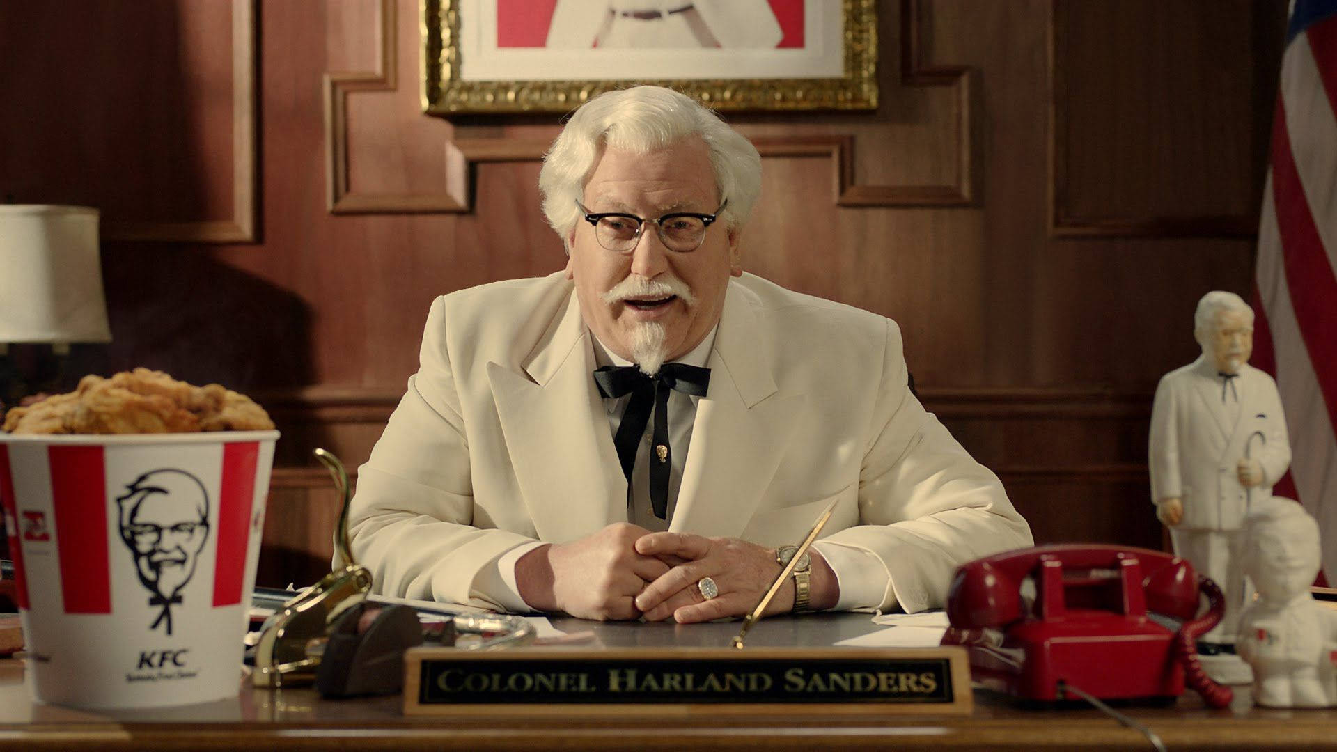 KFC Darrell Hammond 壁紙 — 哈蘭·山德士上校身穿白色西裝打著領結，坐在他的木桌前，周圍環繞著KFC紀念品和一部紅色轉盤電話。