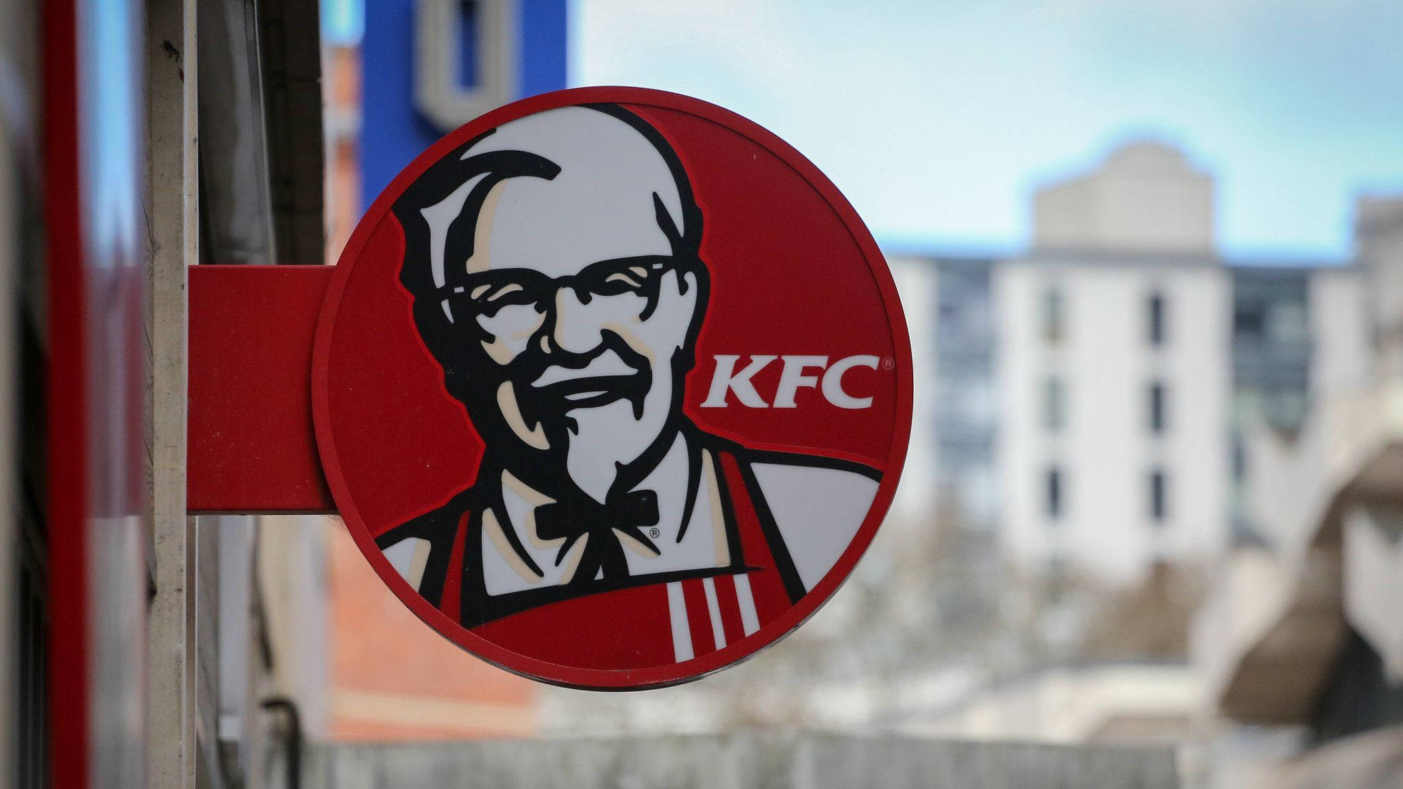 KFC建築招牌 壁紙 — 特寫一個紅色圓形KFC招牌，上面有山德士上校的笑臉，背景是模糊的城市景觀。