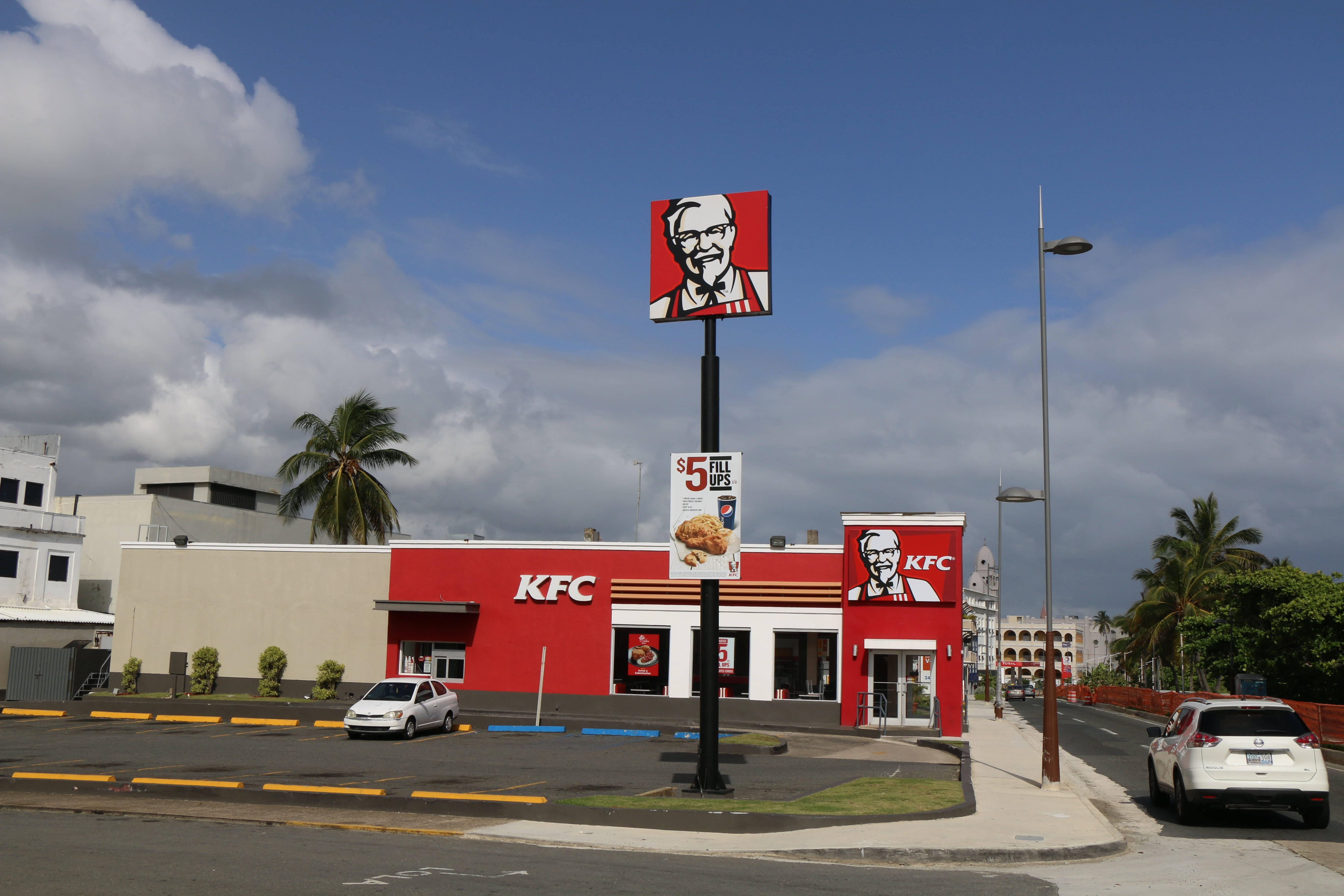KFC快餐店 壁紙 — 一間亮紅色的KFC餐廳，天空部分多雲，有棕櫚樹和停車場的汽車。
