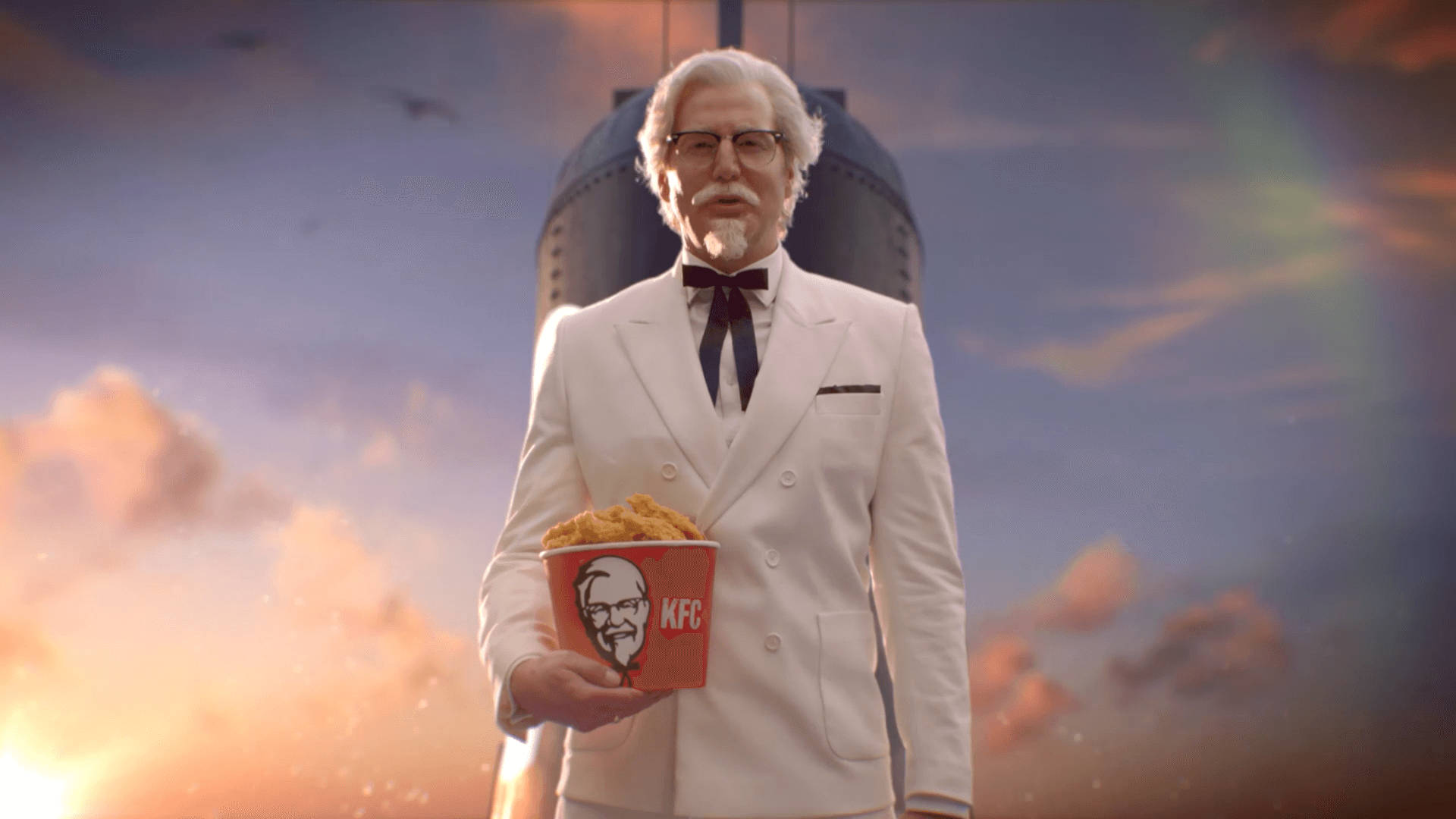 KFC Rob Riggle飾Colonel 壁紙 — 身穿白色西裝的桑德斯上校手持KFC桶，背景是戲劇性的日落天空和火箭。