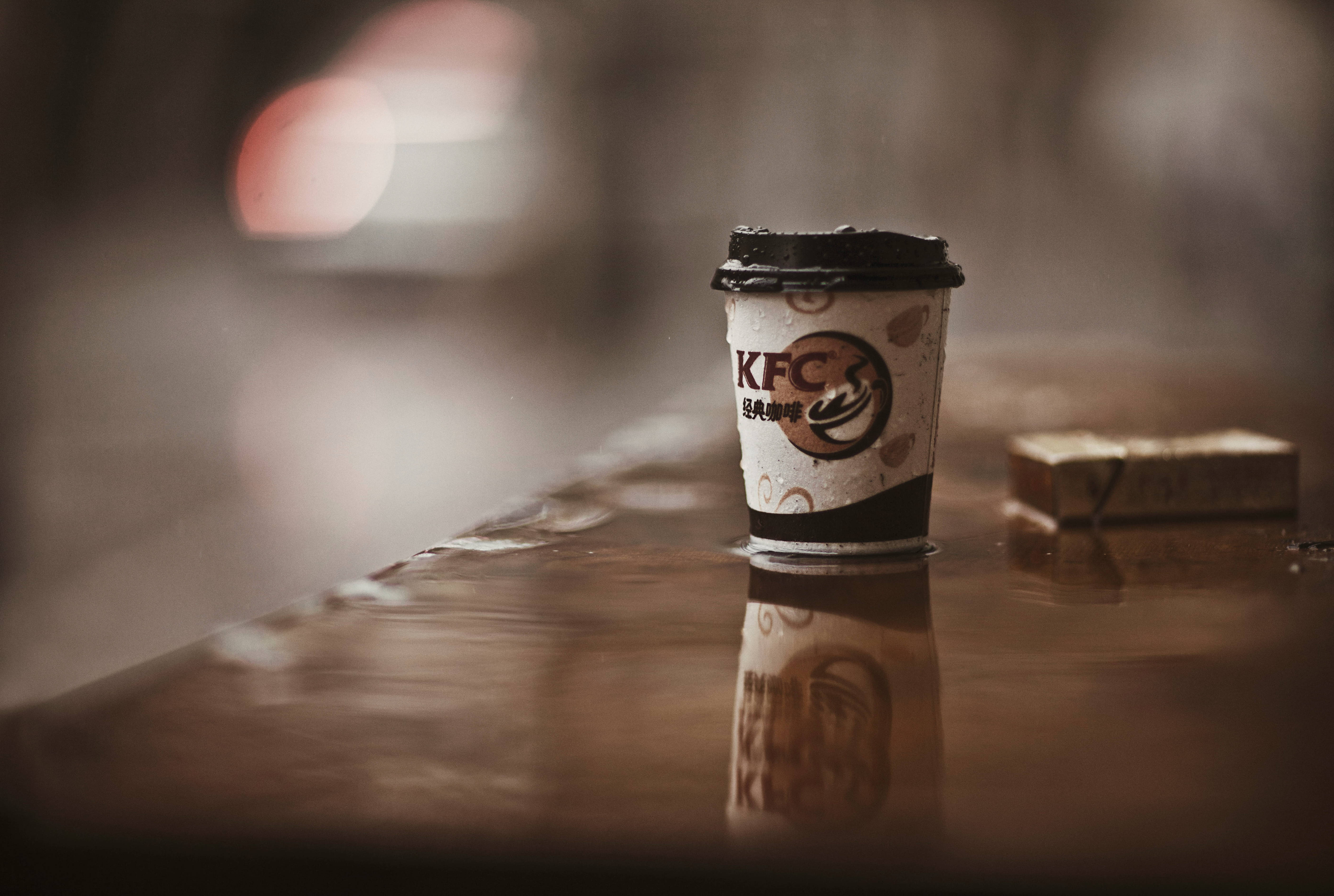 KFC紙杯攝影 壁紙 — 一個被丟棄的KFC咖啡杯放在潮濕反光的桌子上，背景是模糊的城市燈光。