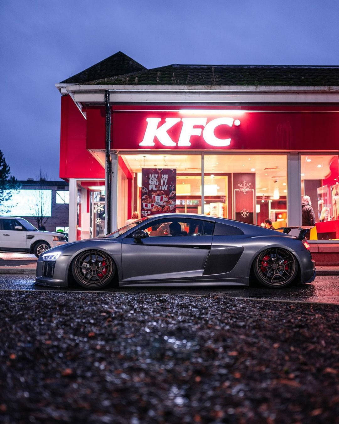 KFC快餐連鎖店 壁紙 — 一輛時尚的銀色跑車在黃昏時停在一間燈火通明的KFC餐廳前，潮濕的路面反射著霓虹燈光。