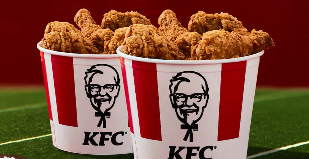KFC 的經典桶裝餐點 壁紙 — 兩個裝滿金黃炸雞的KFC桶，溢出在綠色足球場上，背景為深紅色。