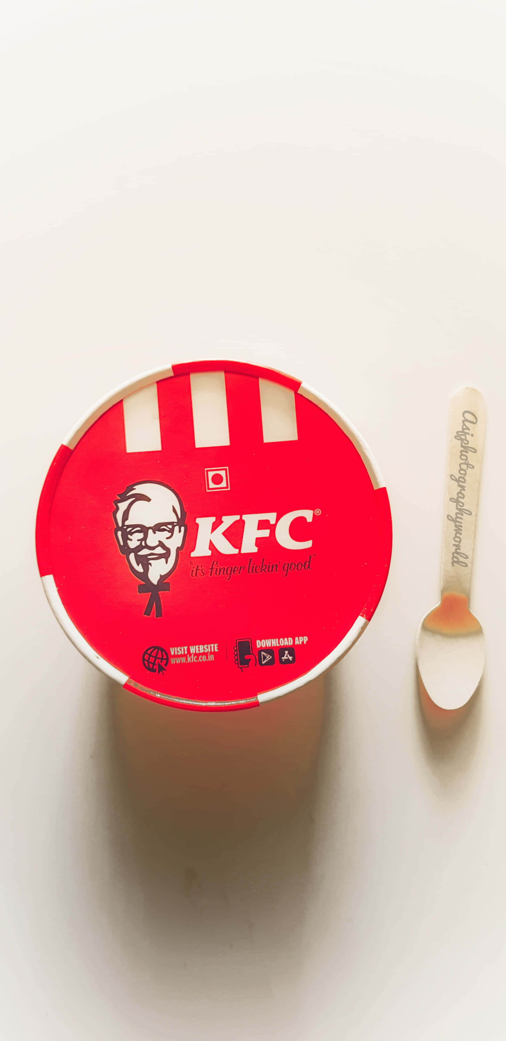 KFC 的美味炸雞 壁紙 — 俯視圖：一個紅色KFC桶，旁邊放著一隻品牌匙羹，置於純白表面上。