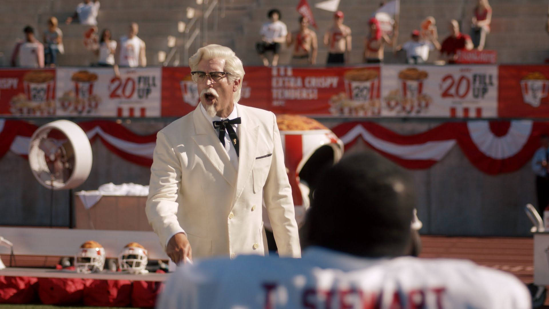 KFC廣告Rob Riggle 壁紙 — 身穿白色西裝的桑德斯上校在陽光明媚的球場上，對著一名足球運動員大喊，場地有紅白旗幟。