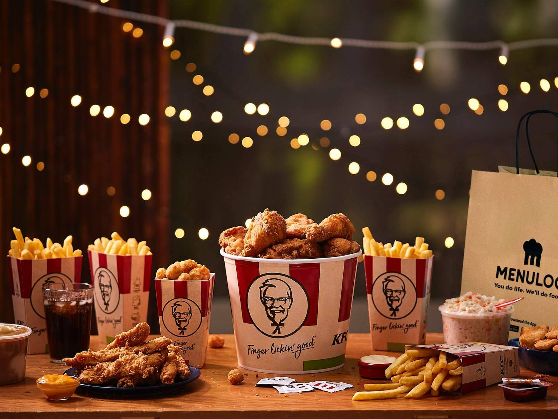 KFC盛宴套餐 壁紙 — 夜晚，溫暖的串燈下，一張木桌上擺滿了KFC炸雞桶、薯條和飲料。