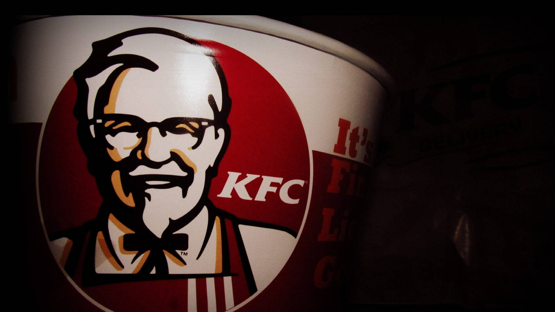 KFC雞桶聚焦攝影 壁紙 — KFC 桶的近距離特寫，在戲劇性的低光照明下展示山德士上校的標誌性標誌。