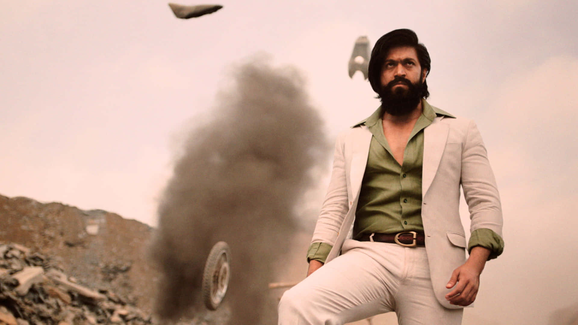 《KGF 2》劇照：羅基·巴伊眼神凌厲，背景為科拉金礦區 壁紙 — 一位留鬍鬚的男人身穿白色西裝，自信地站在煙霧和殘骸中，天空中有飛行車輛。