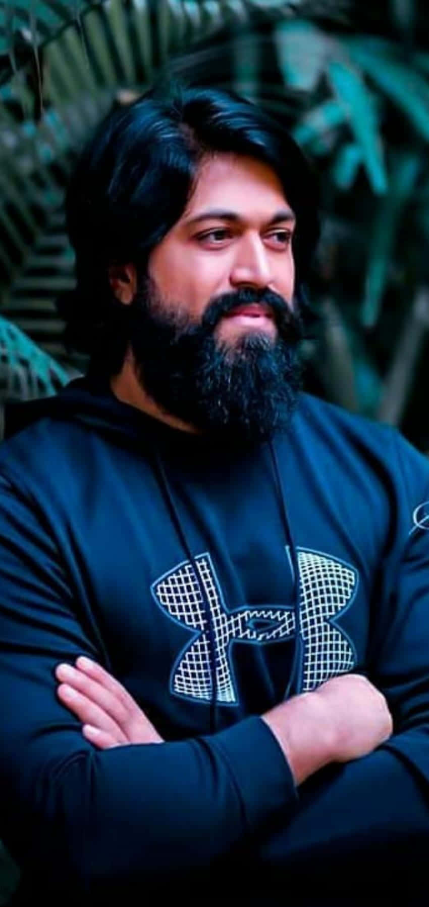一位留鬍子的男人穿著黑色連帽衫，在綠色戶外環境中交叉雙臂站立。