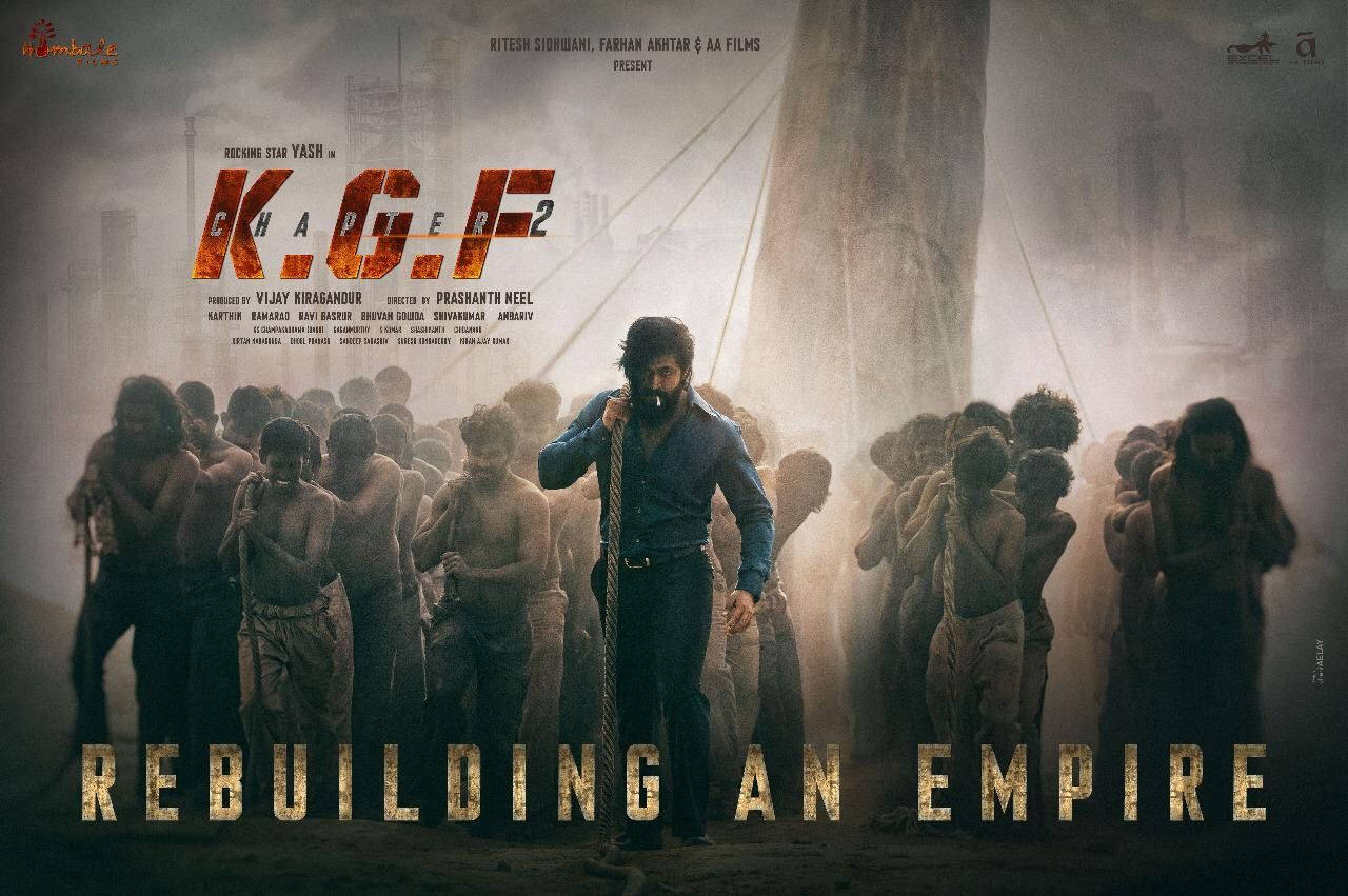 KGF Chapter 2 羅基與奴隸 壁紙 — 一張粗獷的電影海報顯示一個鬍鬚男人穿著藍色襯衫拿著繩子，周圍是赤裸上身的男人，在塵土飛揚的城市環境中，光線戲劇化。
