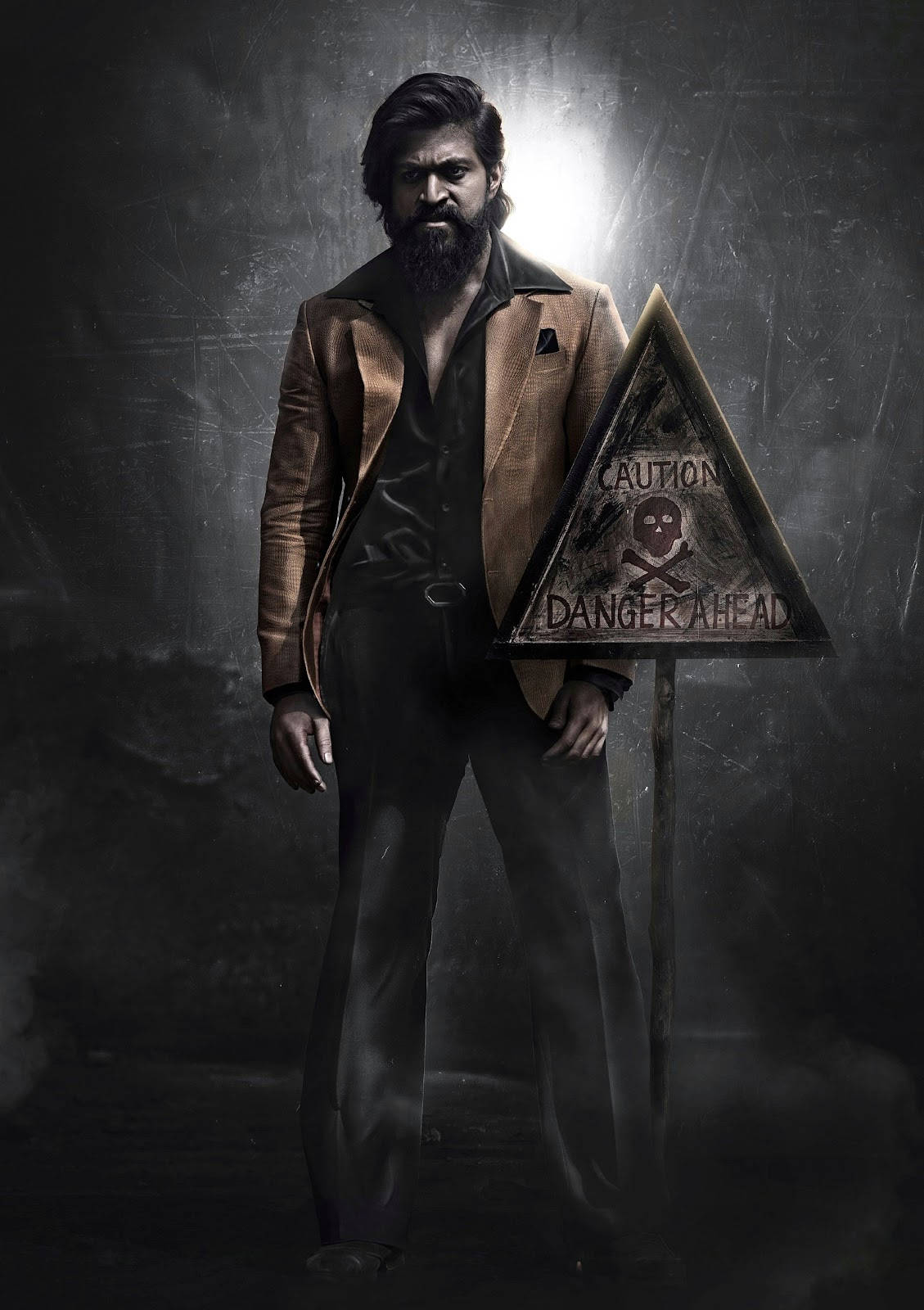 KGF Rocky Bhai 站在危險標誌前 壁紙 — 一個留鬍鬚的男人穿著棕色外套，在戲劇性的聚光燈下威脅性地站在「危險 ahead」警告標誌旁邊。
