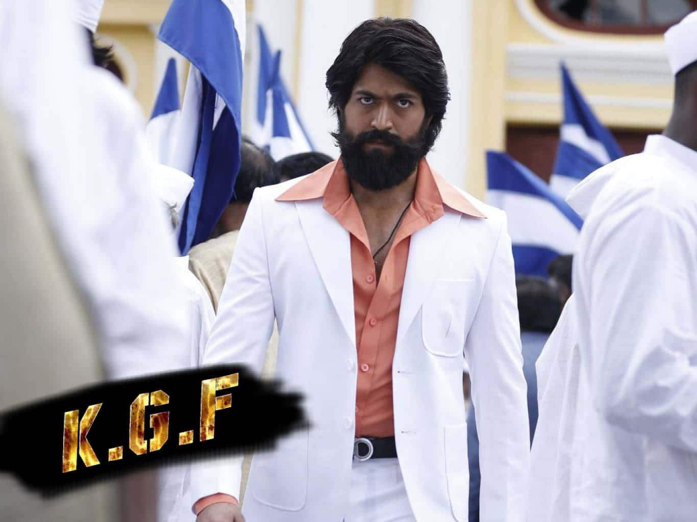 KGF - 鋼鐵披甲的失落王國之夢 壁紙 — 一位留鬍鬚的男子身穿白色西裝和橙色襯衫，自信地站在手持藍白旗幟的人群中。