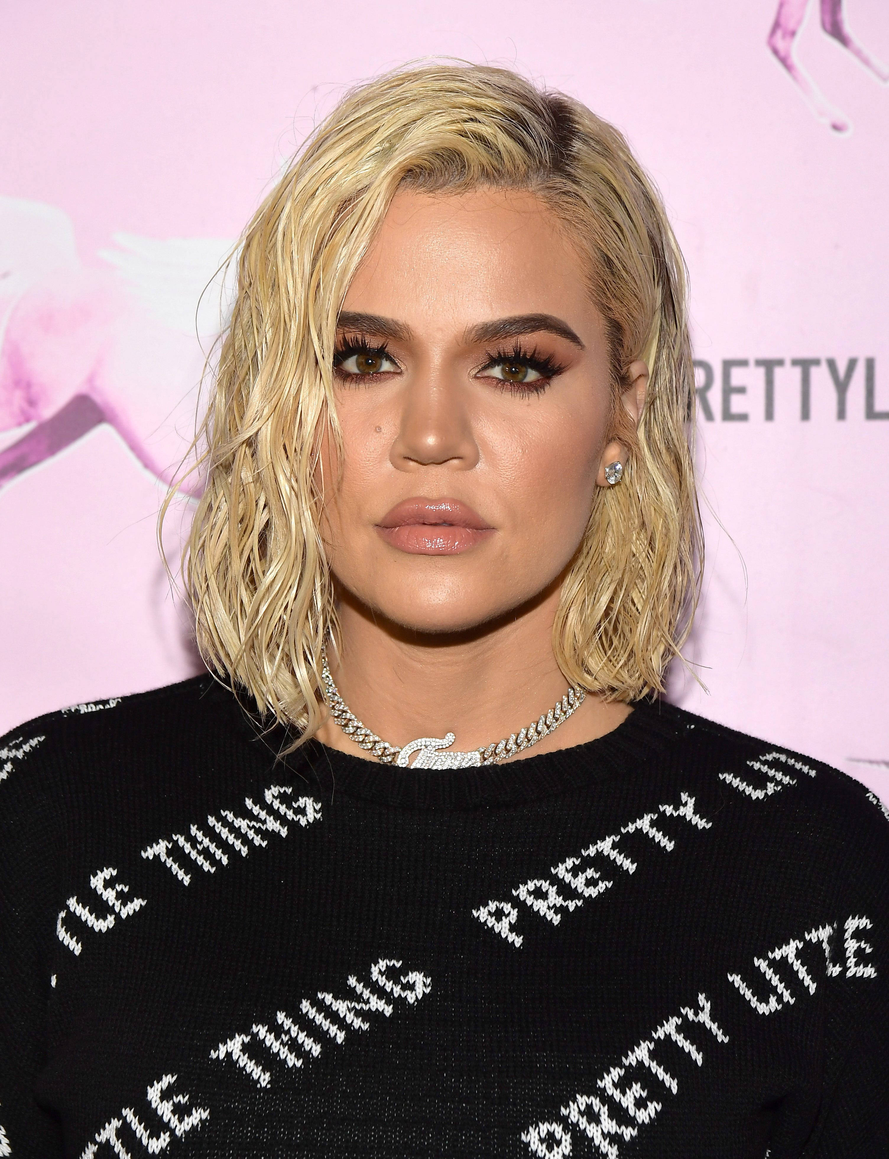 Khloe Kardashian 嘴脣豐盈特寫 壁紙 — Khloé Kardashian 在粉紅色品牌背景前穿著帶有白色文字的黑色毛衣擺姿勢。