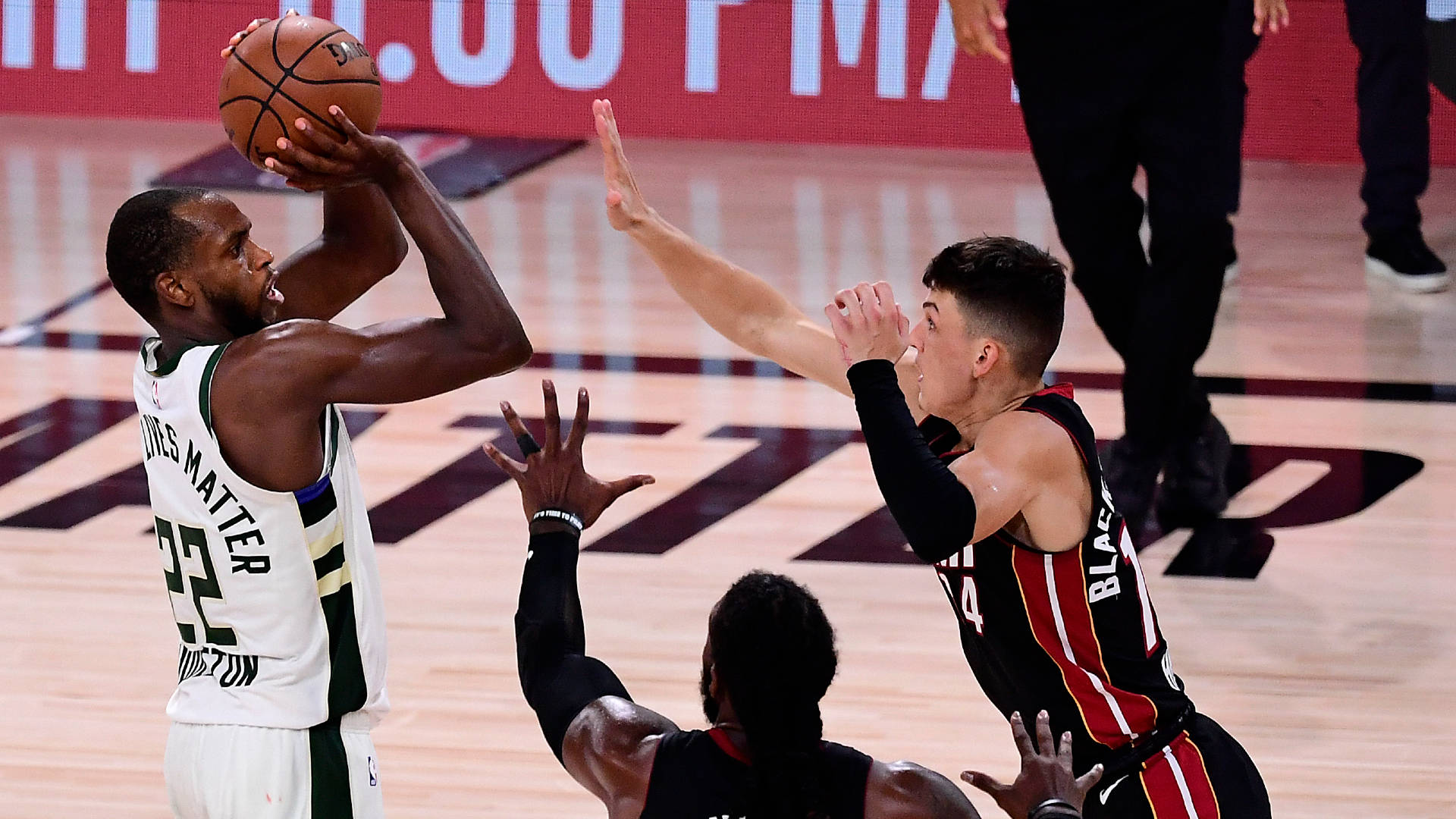 Khris Middleton 與 Tyler Herro 壁紙 — 一名身穿白色球衣的籃球運動員在燈光明亮的球場上，在對手的防守下投籃。