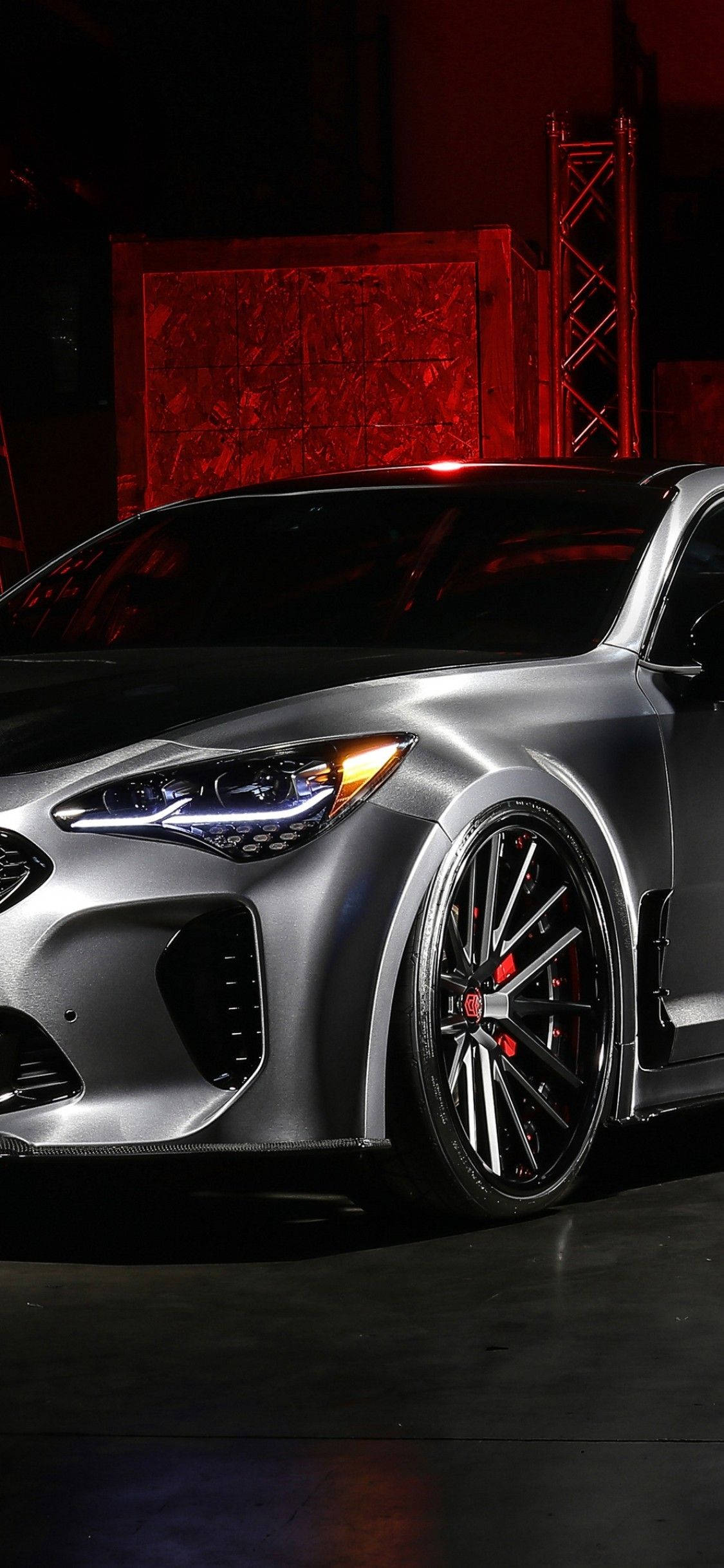 高雅的Kia Stinger 壁紙 — 一輛時尚的銀色跑車，帶有發光的頭燈和紅色制動卡鉗，在黑暗的工業車庫中，在戲劇性的紅色燈光下展示。