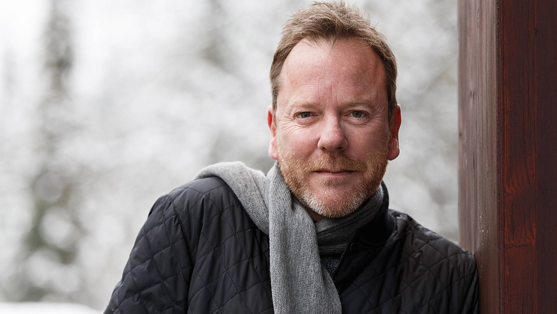 Kiefer Sutherland 在加拿大惠斯勒村 壁紙 — 一個留鬍子、戴圍巾的男人靠著木門，身處白雪皚皚的冬日場景。