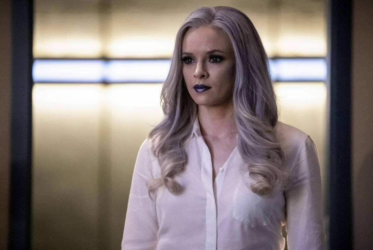 Killer Frost 穿白色上衣 壁紙 — 一位銀色頭髮、深色唇膏的女性站在光線柔和的走廊中，身後有發光的水平面板。