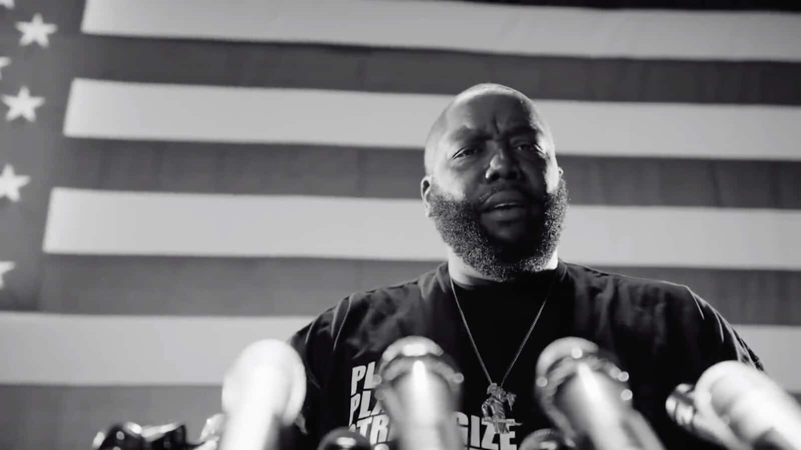 Killer Mike 在講台演講 壁紙 — 一位鬍鬚男在記者會上發言的黑白肖像，背景有麥克風和美國國旗。