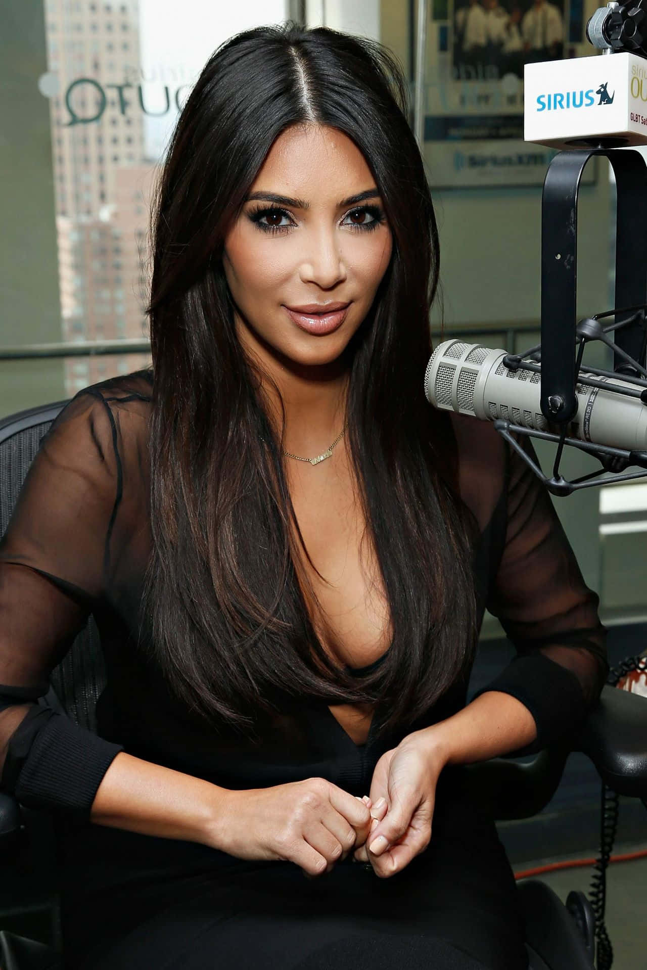 金·卡戴珊 桌布 壁紙 — Kim Kardashian 在攝影棚內從容就座，身穿黑色透視上衣，附近有麥克風。