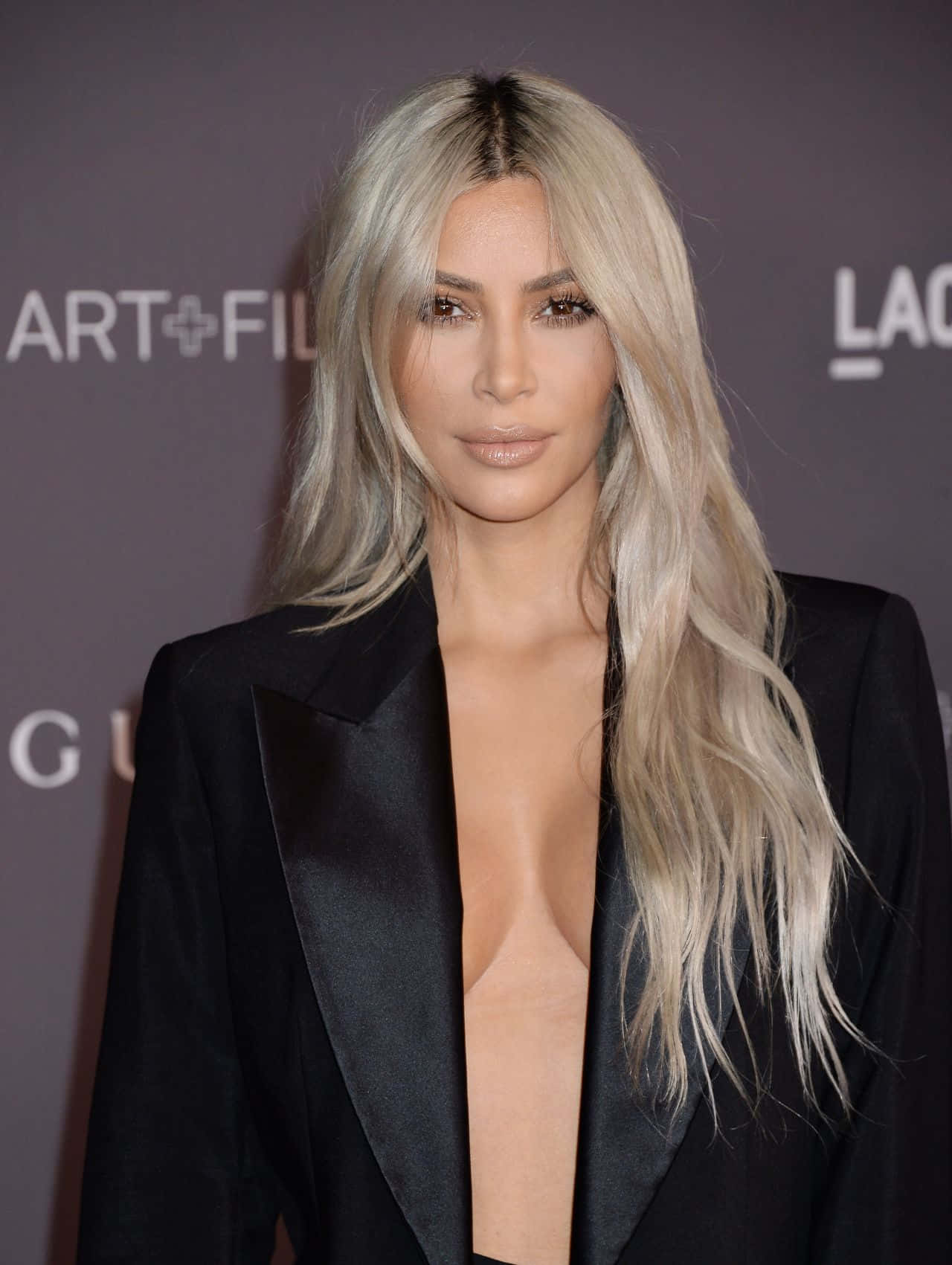 金·卡戴珊 桌布 壁紙 — Kim Kardashian 在品牌活動背景前，身穿時尚黑色西裝外套，留著長白金髮，擺出姿勢。
