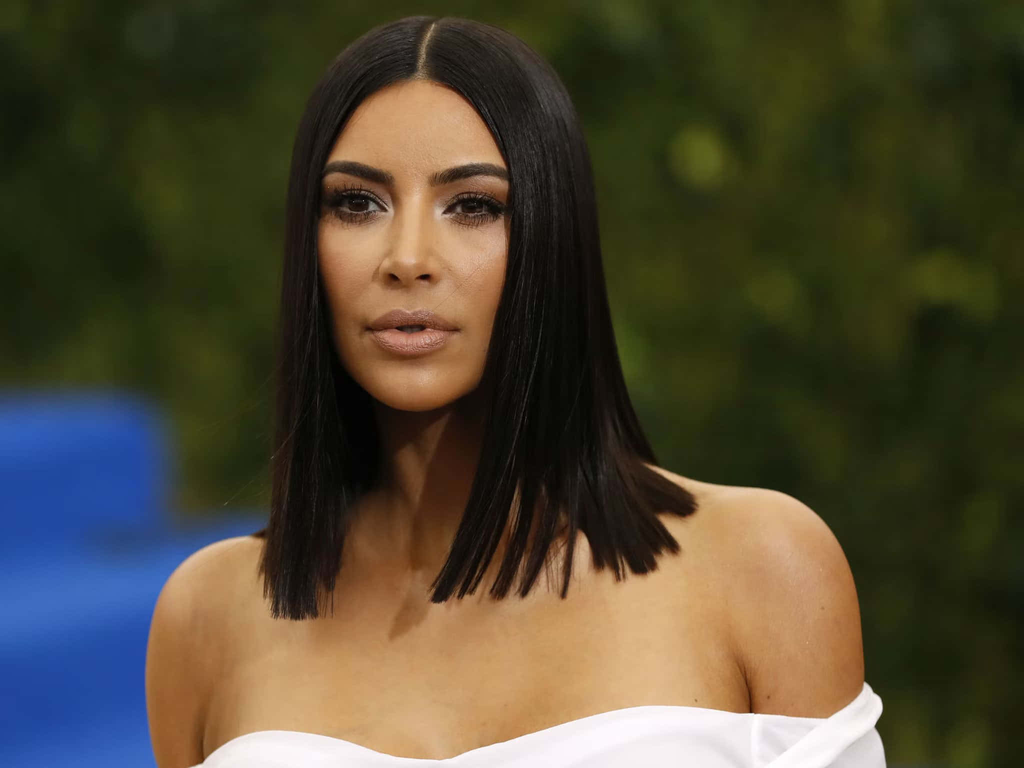 金·卡戴珊 桌布 壁紙 — Kim Kardashian 穿著白色露肩連衣裙，搭配光滑的黑色鮑伯頭，在模糊的綠色和藍色背景前擺姿勢。