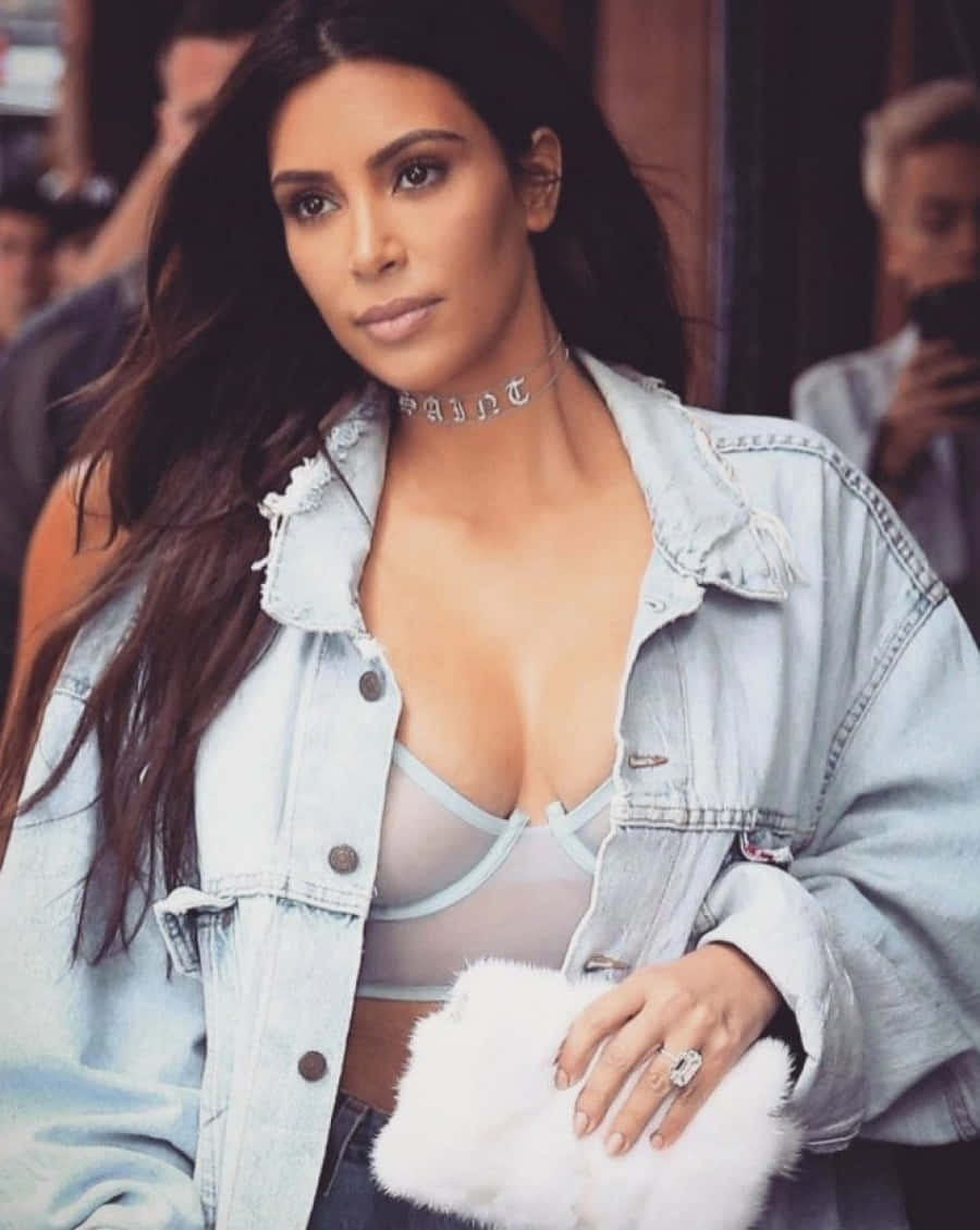 金·卡戴珊 桌布 壁紙 — Kim Kardashian 自信地擺姿勢，身穿淺色牛仔外套內搭透視上衣，手持白色毛絨手拿包。