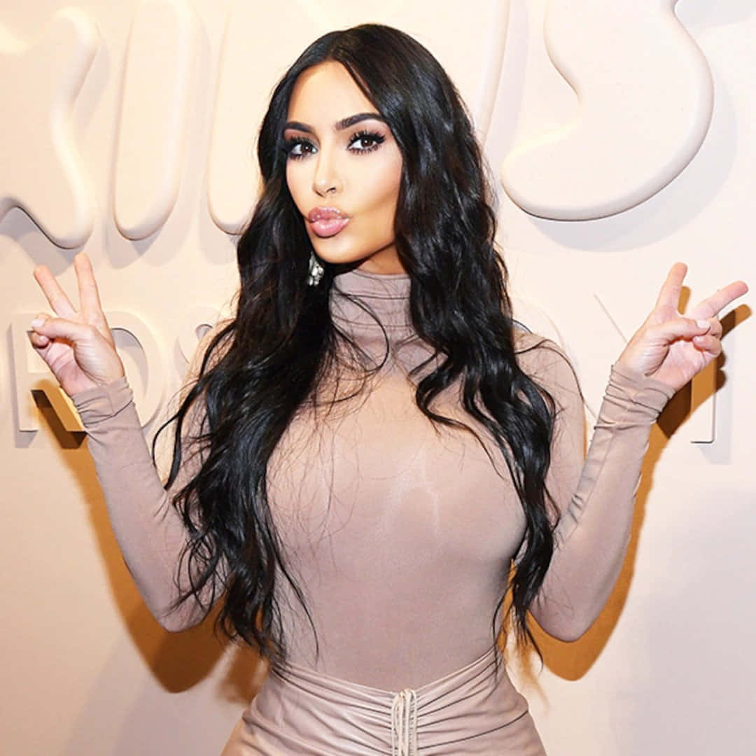 金·卡戴珊 桌布 壁紙 — Kim Kardashian 自信地擺姿勢，身穿透明米色連體衣，對著白色品牌牆，雙手比出和平手勢。