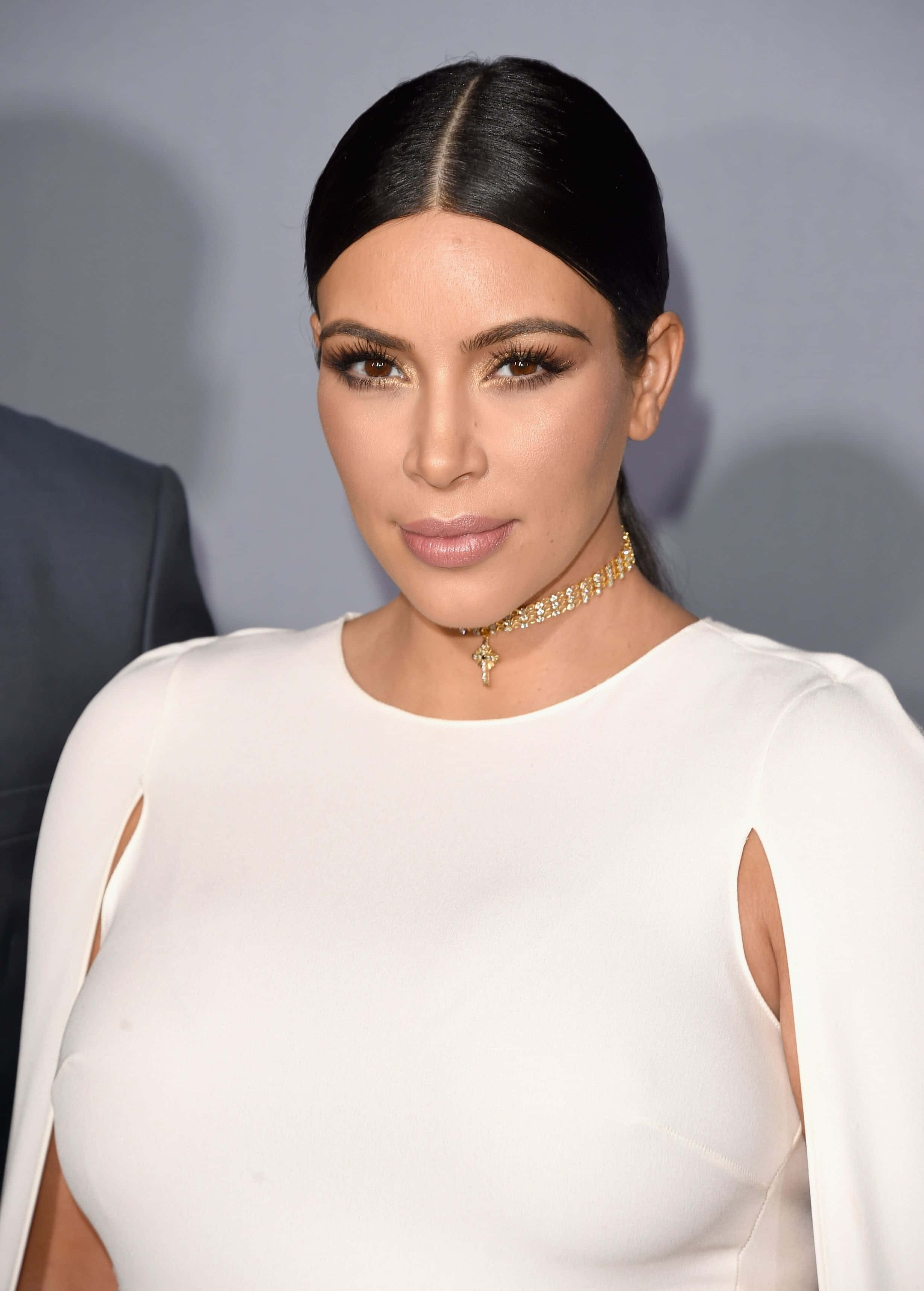 Kim Kardashian 桌布 壁紙 — Kim Kardashian 身穿一襲帶有鏤空設計的修身白色連衣裙，搭配金色頸鏈，在柔和的灰色背景前擺姿勢。