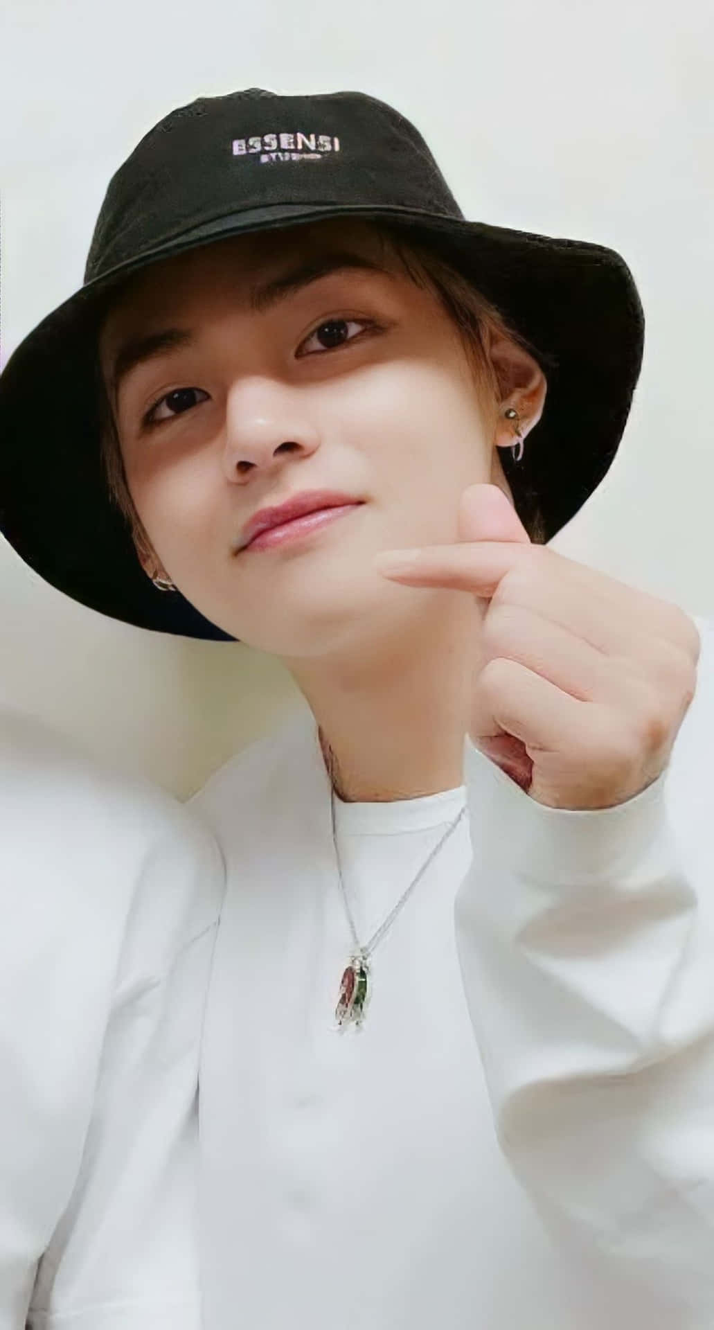 Kim Taehyung BTS 指心 壁紙 — 一位戴黑色漁夫帽、穿白色襯衫的年輕男子，在純色背景前微笑指著自己的臉。