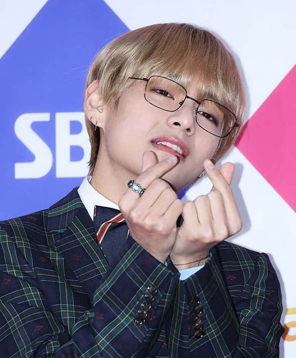 Kim Taehyung BTS 指心 壁紙 — 一位金髮戴眼鏡的年輕男子，穿著格紋西裝，在色彩繽紛的幾何背景前做出心形手勢。