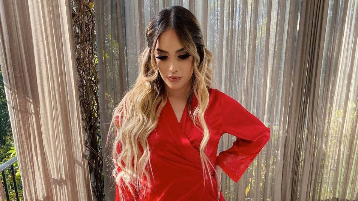 墨西哥歌手Kimberly Loaiza身穿紅袍 壁紙 — 一位身穿鮮紅色緞面連衣裙的女性在陽光穿透的薄紗窗簾旁自信地擺姿勢。