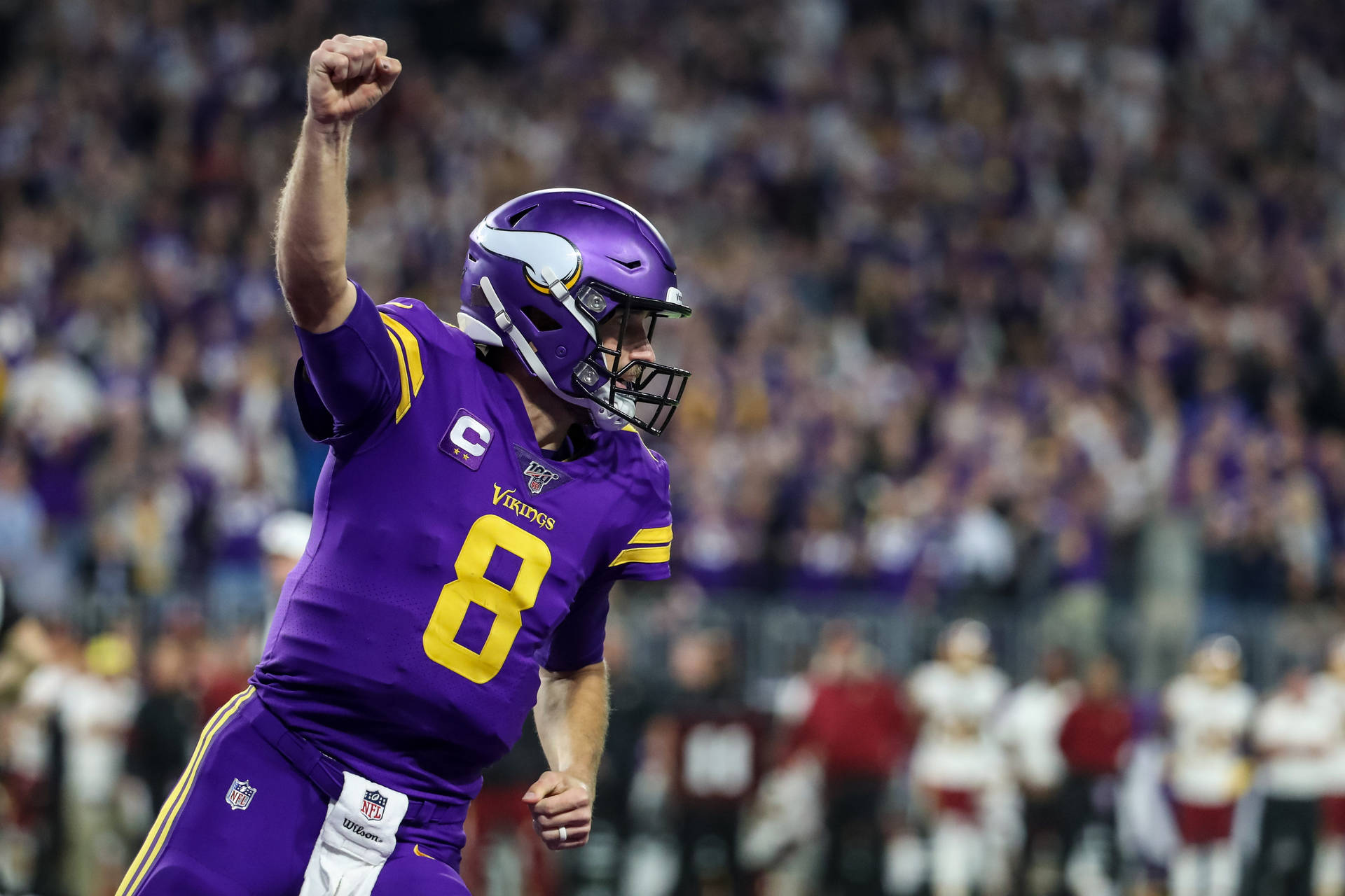 Kirk Cousins 維京人隊握拳慶祝 壁紙 — 明尼蘇達維京人隊的四分衛身穿紫色和金色球衣，在夜間比賽中於球場上舉起拳頭慶祝勝利。
