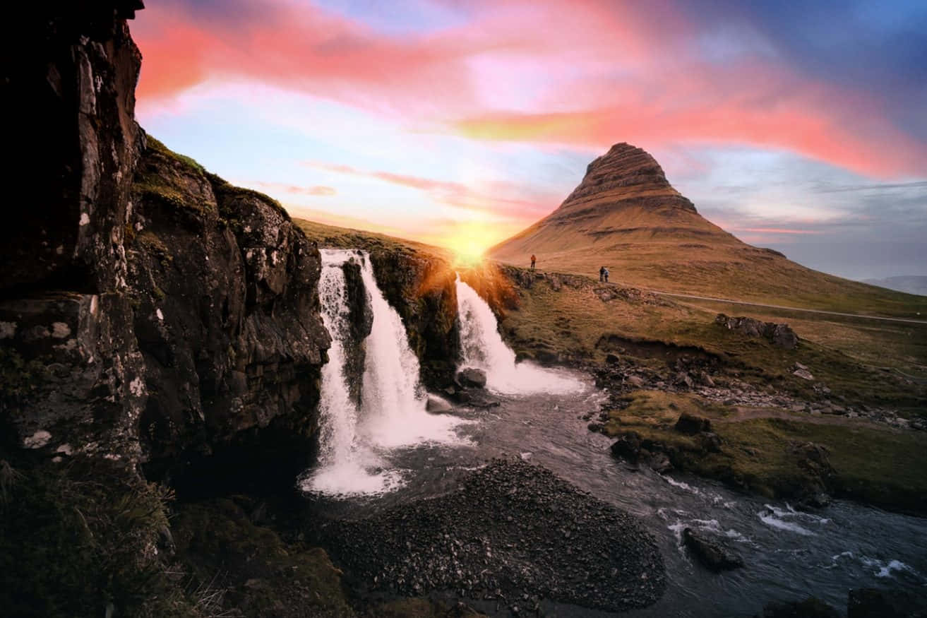 Kirkjufell 山與夢幻粉彩天空 壁紙 — 一道雄偉的瀑布在熾熱的日落天空下傾瀉入岩石溪流，遠處有一座孤獨的山峰。