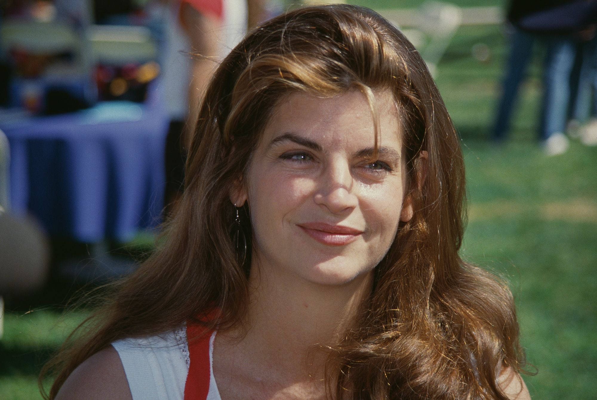 年輕的 Kirstie Alley 參加加州名人壘球賽 壁紙 — 一位長棕髮女性在陽光明媚的日子裡於戶外溫暖地微笑，背景是模糊的綠草和藍色桌布。